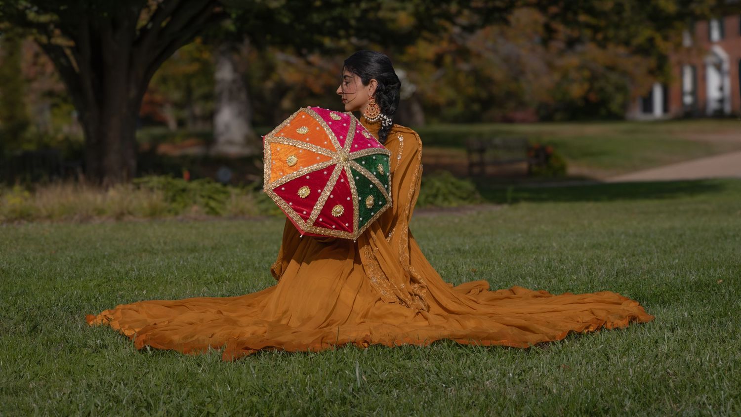Woman in orange lehenga sits on grass holding a colorful embroidered umbrella.