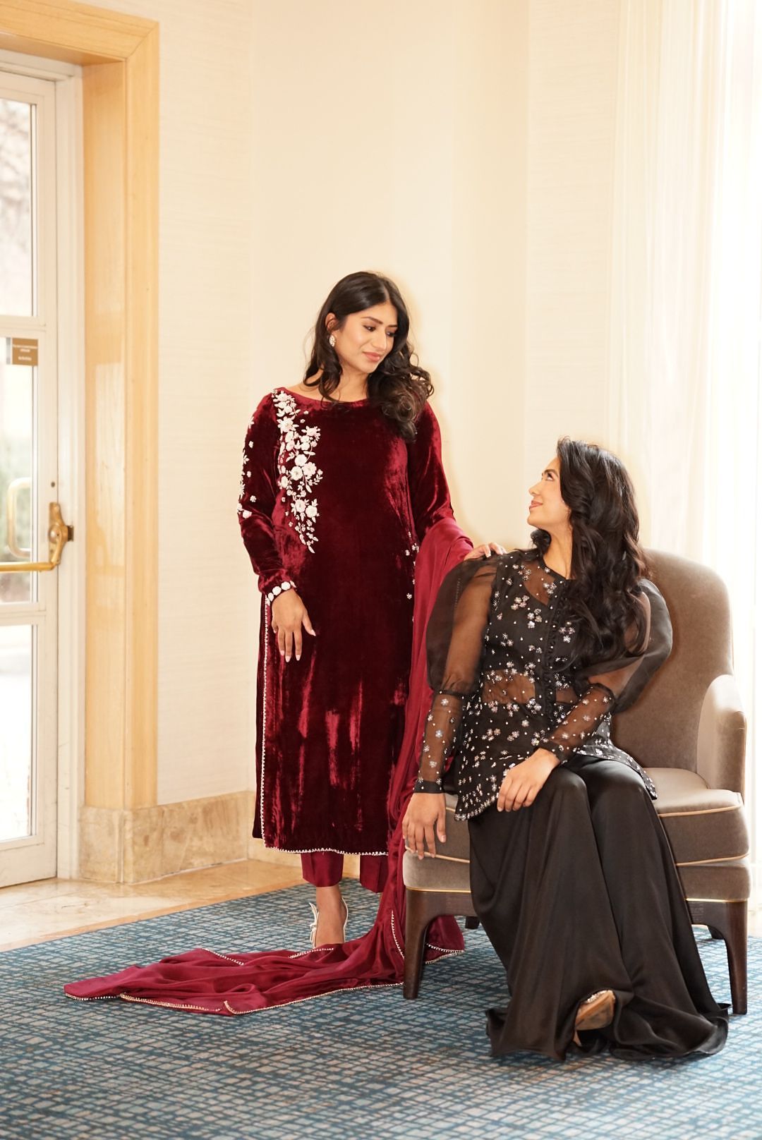 Two women in elegant South Asian attire, one in crimson velvet salwar kameez, one in black embellished outfit.