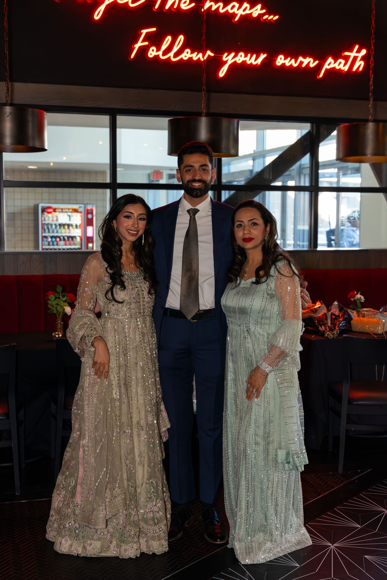 Three people posing in formal South Asian attire at restaurant with neon motivational sign backdrop.