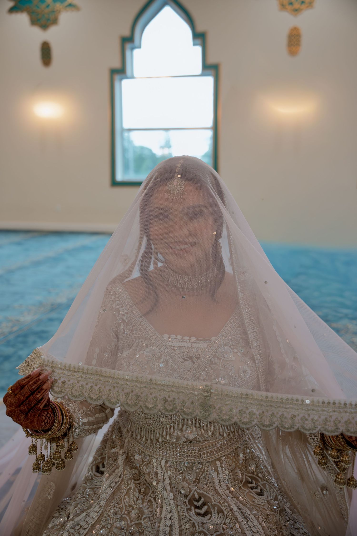 South Asian bride in white lehenga and veil holds dupatta in ornate mosque setting.