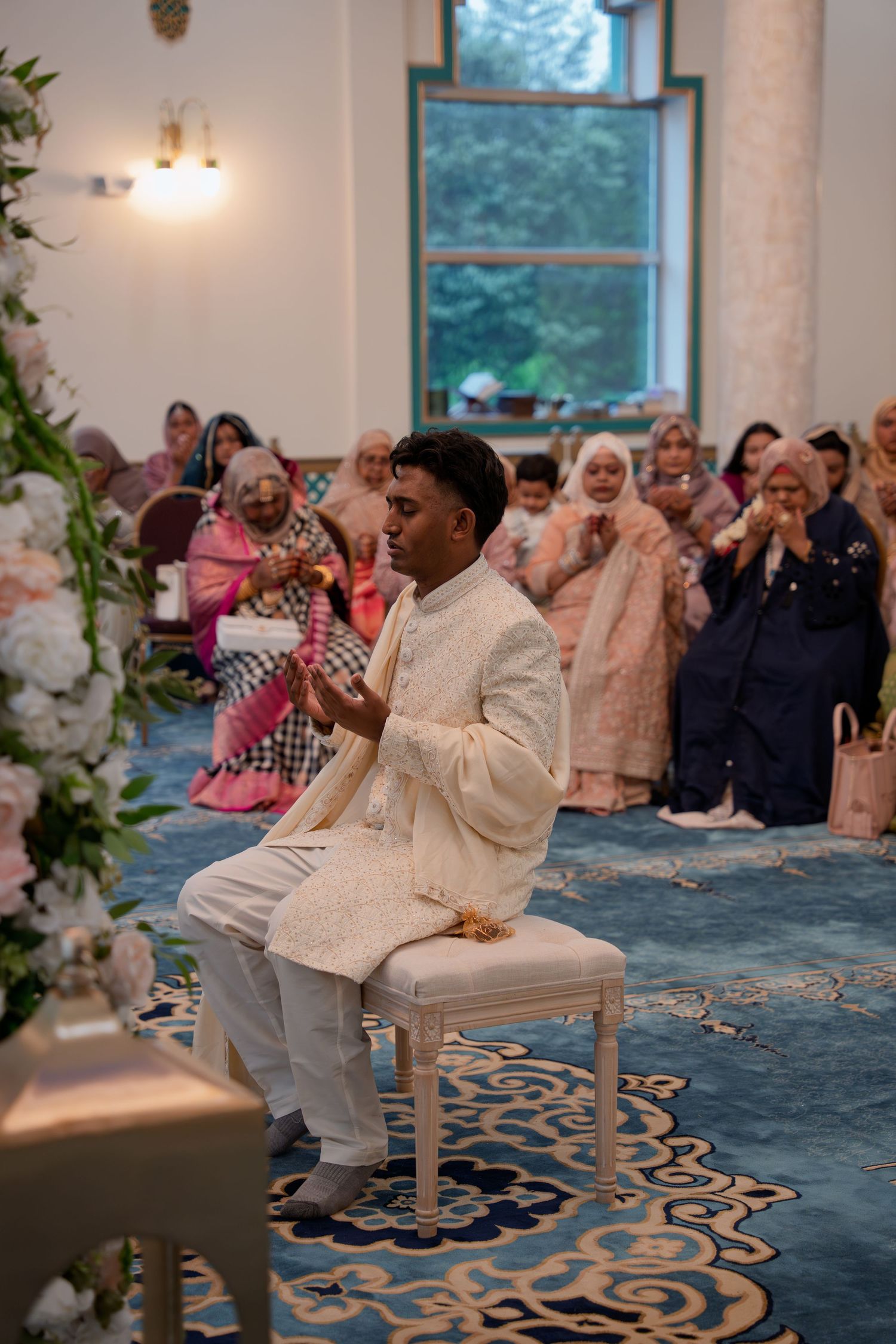 Groom in ivory sherwani sits during Muslim wedding ceremony, guests in traditional attire seated behind him.