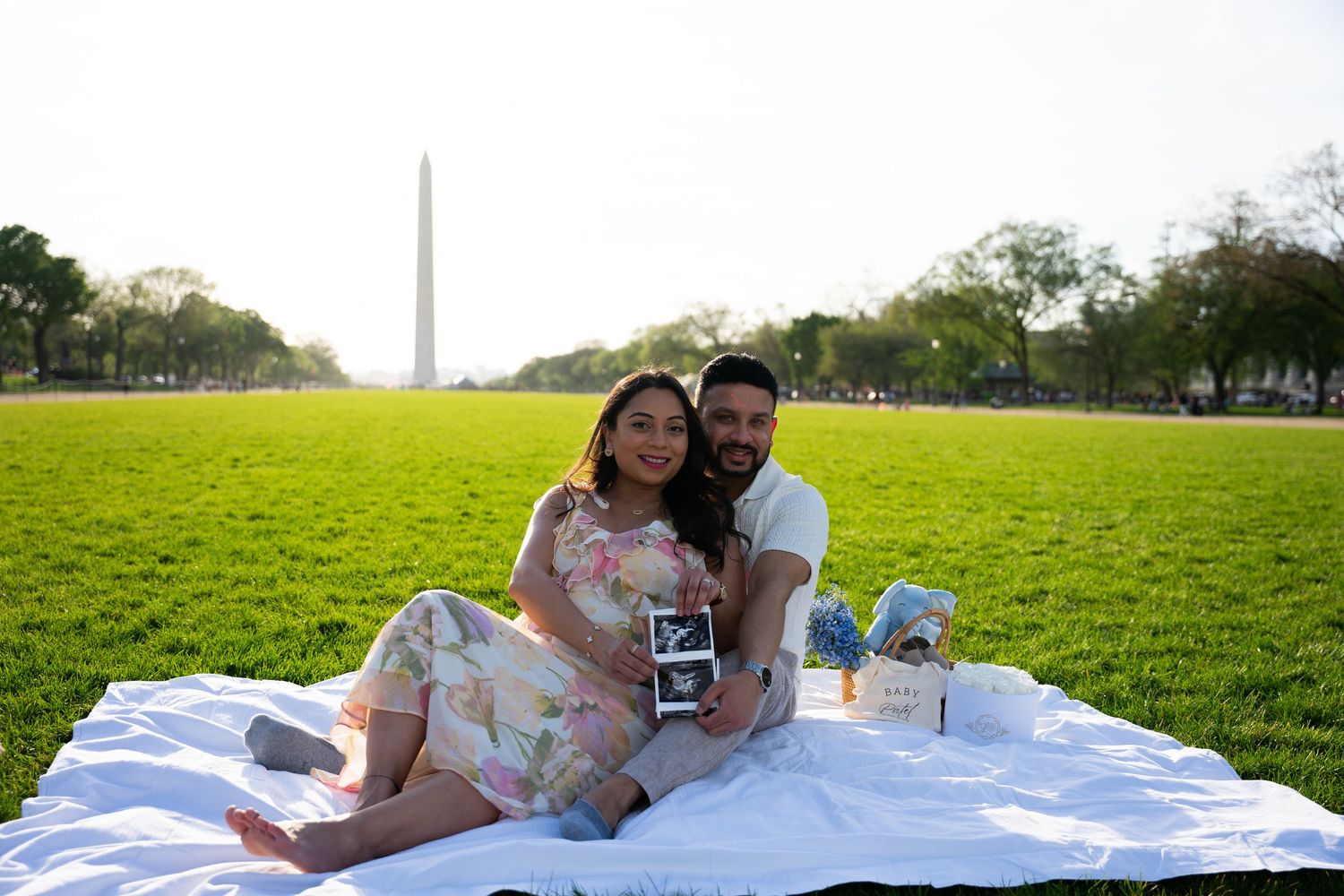 Couple sitting on white blanket at National Mall with Washington Monument backdrop holding ultrasound photo.