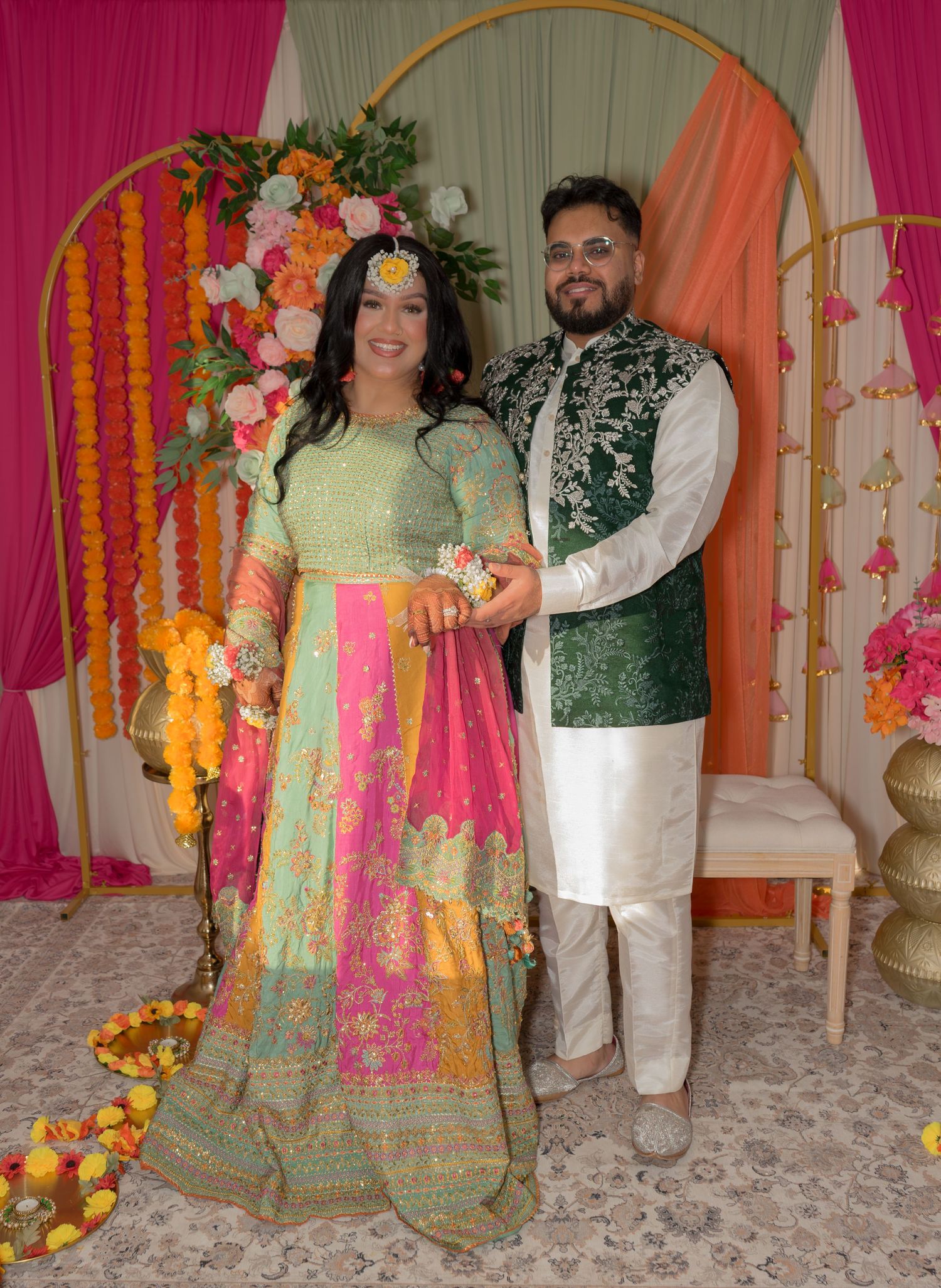 Couple in vibrant traditional South Asian mehndi ceremony attire posing before colorful floral backdrop.