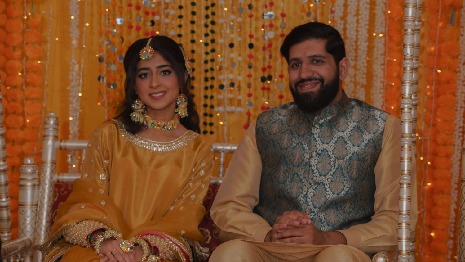 Pakistani couple in traditional golden outfits smiling together at mehndi ceremony with marigold backdrop.