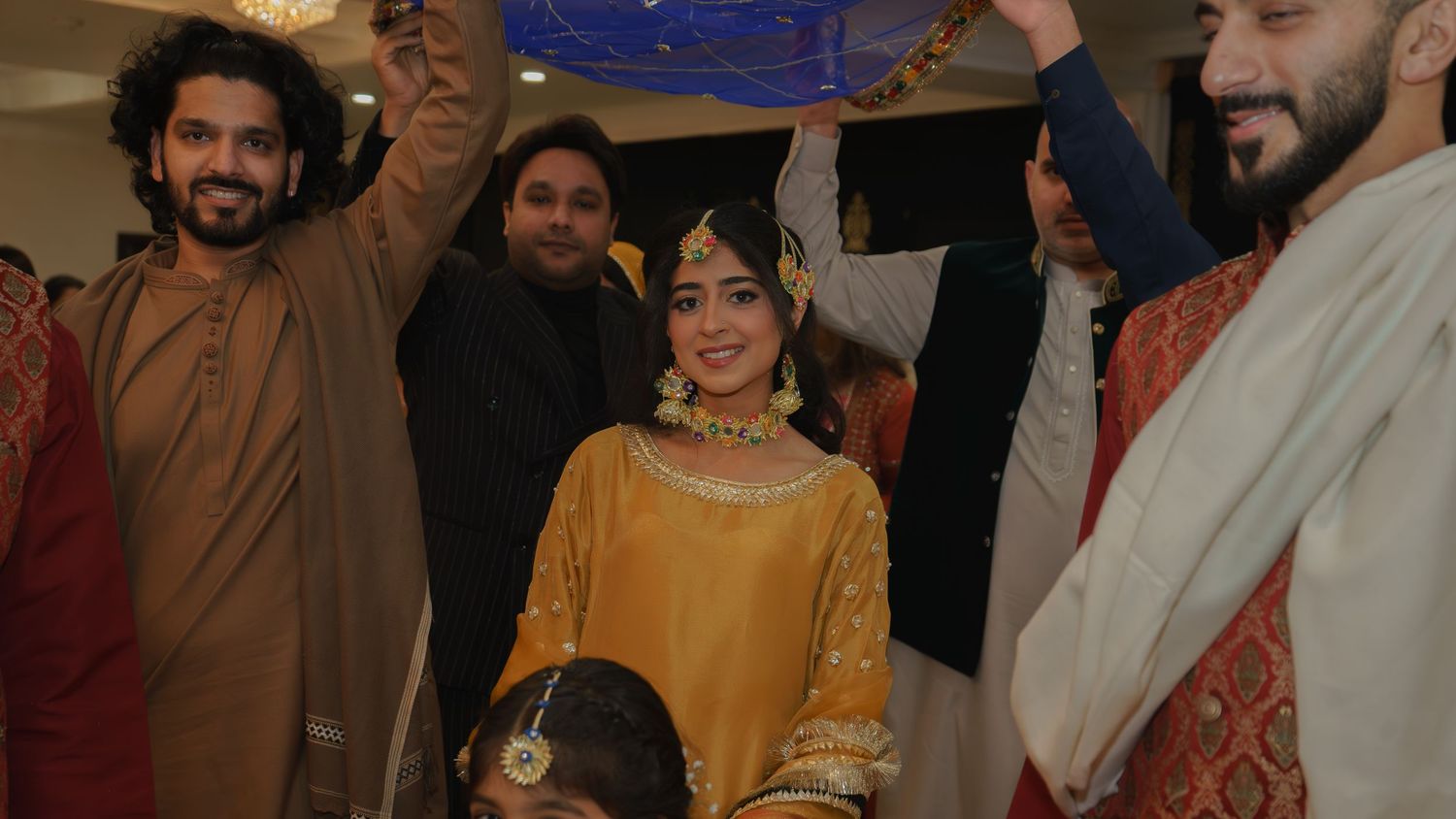A bride in a golden yellow outfit smiles during a traditional South Asian wedding ceremony surrounded by guests.