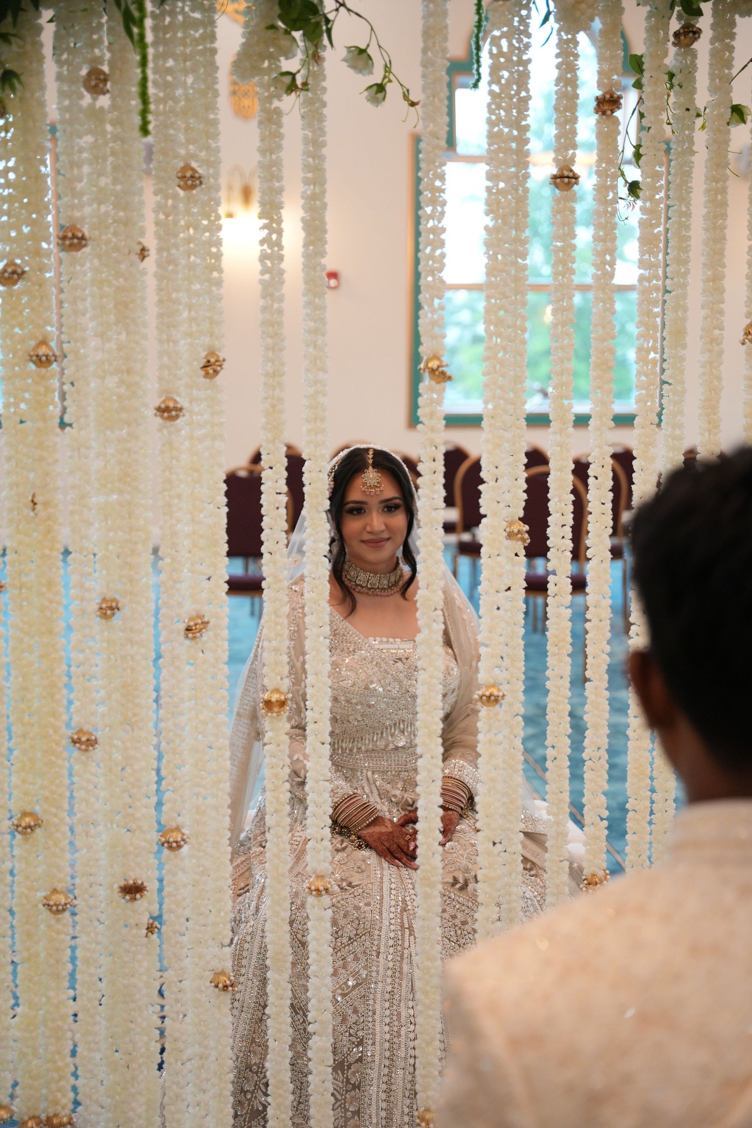 Bride in white embroidered lehenga stands behind floral curtain of jasmine strings at an Indian wedding ceremony.