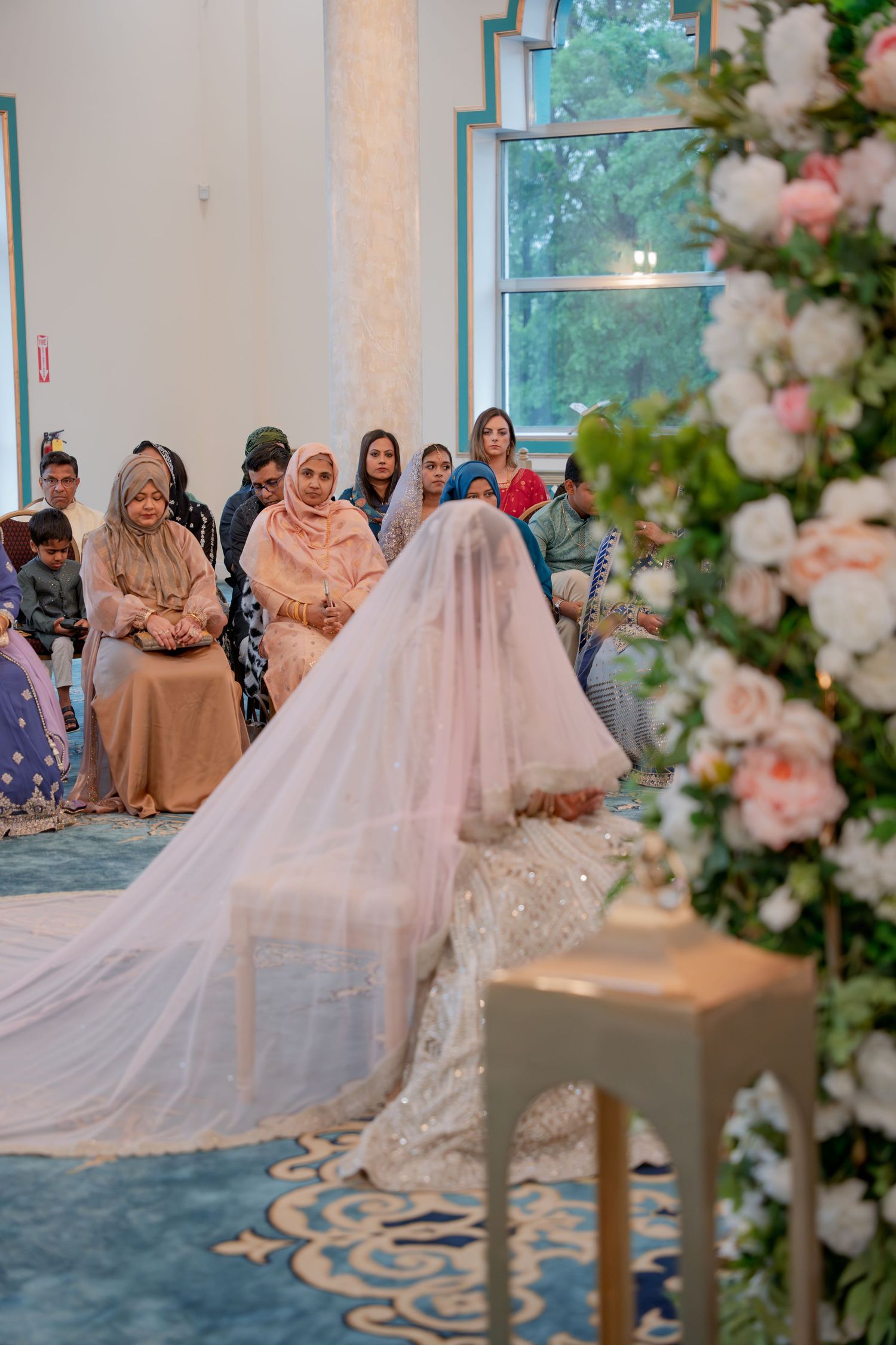 Veiled bride in white lehenga sits before guests during Islamic nikah wedding ceremony in mosque.