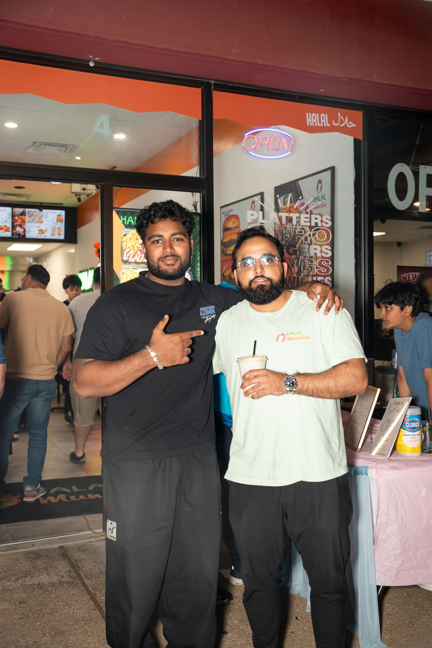 Two men posing outside halal restaurant storefront with crowd in background.