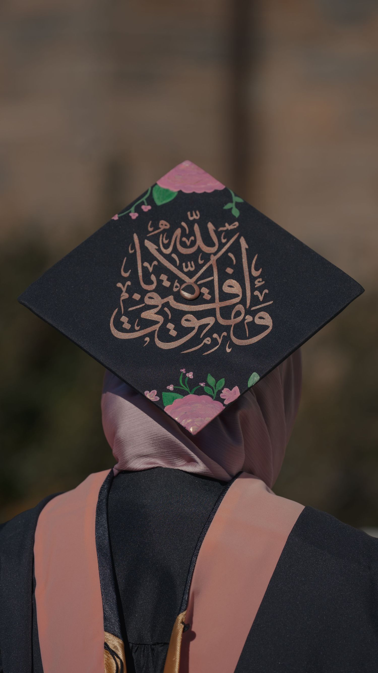 Graduate wearing decorated cap with Arabic calligraphy and floral designs at commencement ceremony.