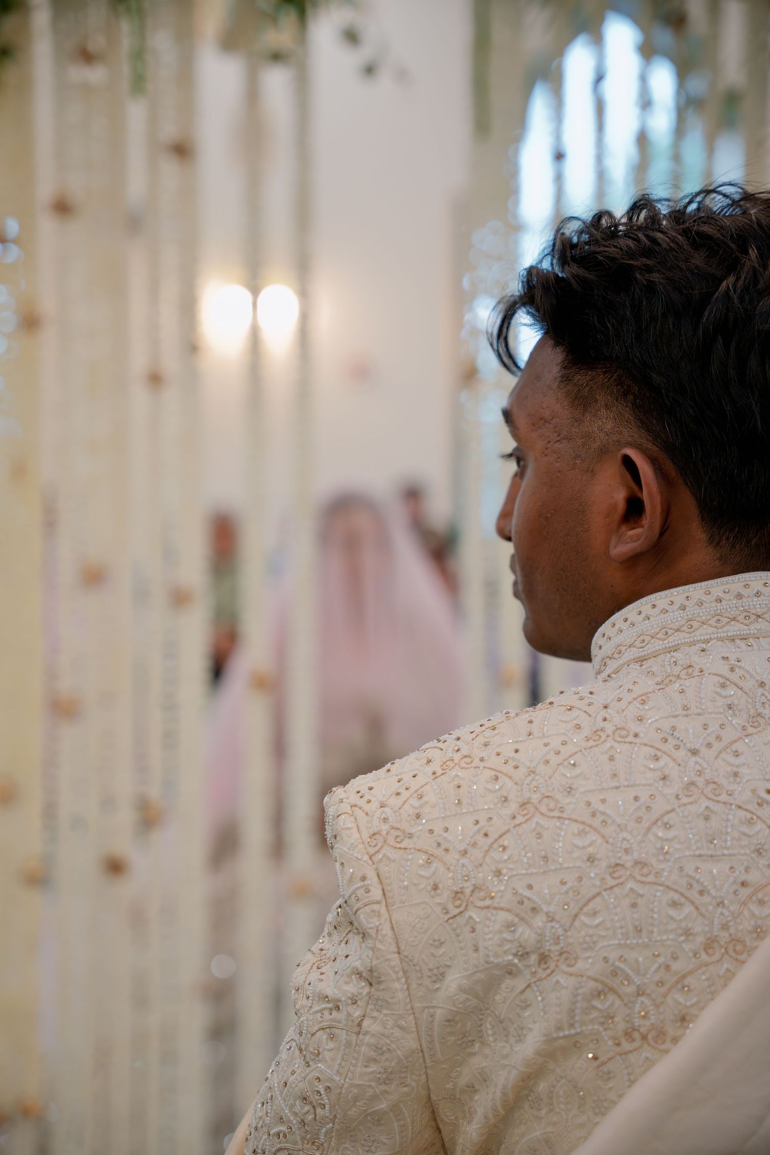 Groom in embroidered sherwani watches bride approach in decorated wedding venue.