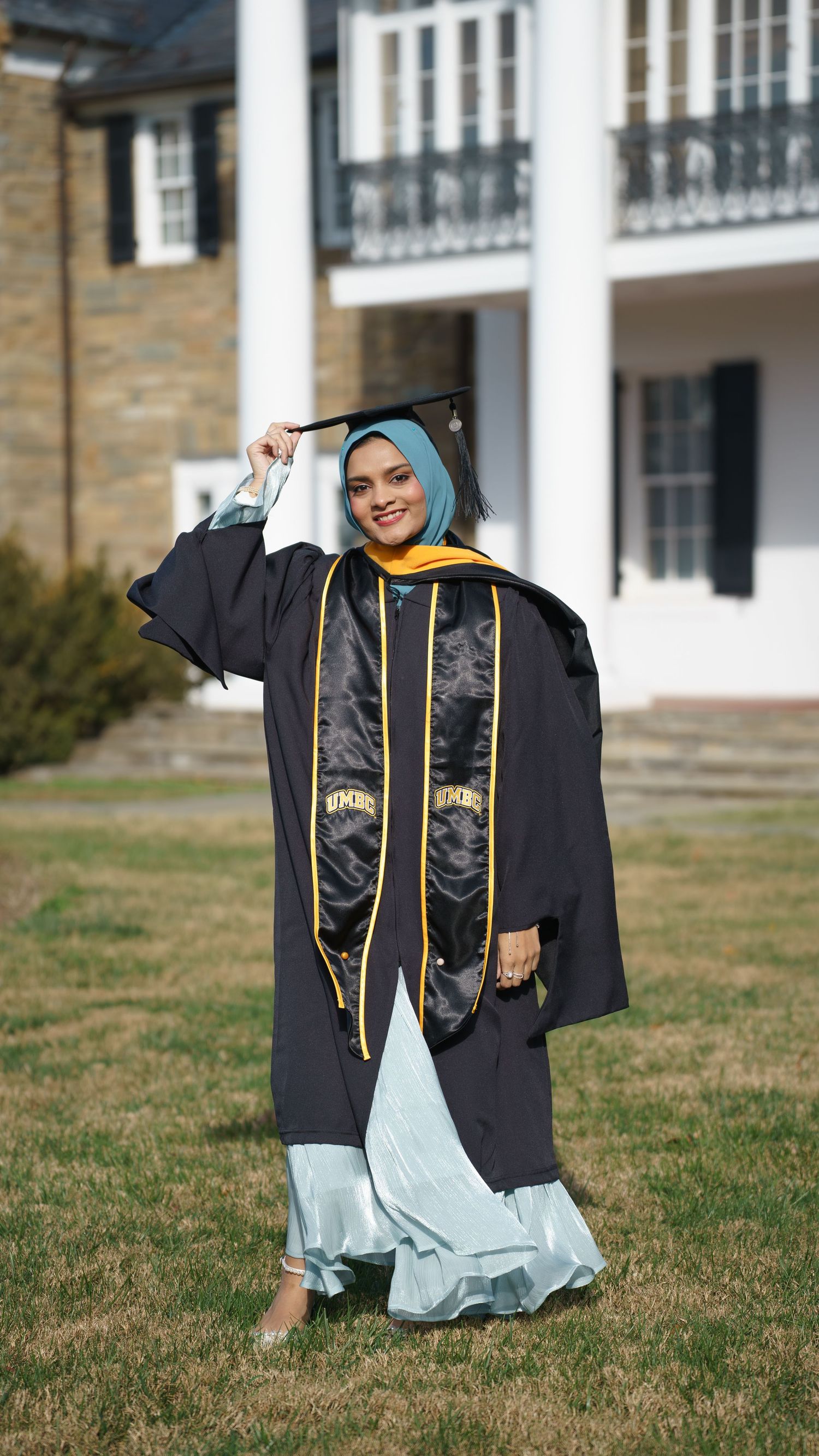 Graduate in black cap and gown with gold hood and hijab posing outdoors at UMBC campus.