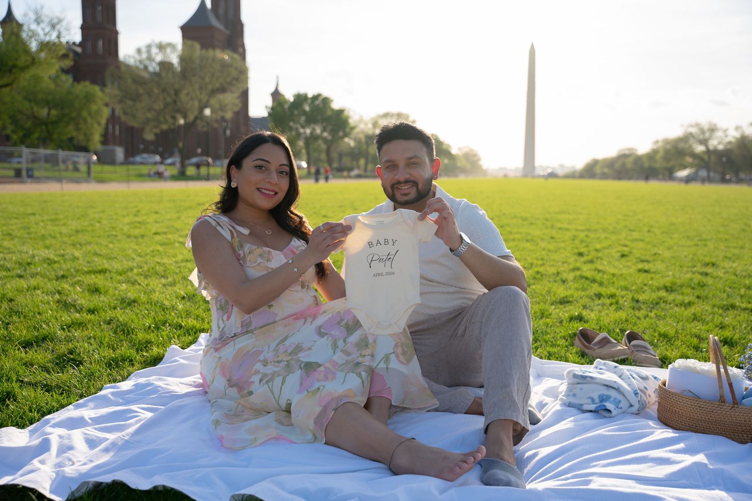 Couple sitting on blanket at National Mall holding baby onesie with Washington Monument in background.
