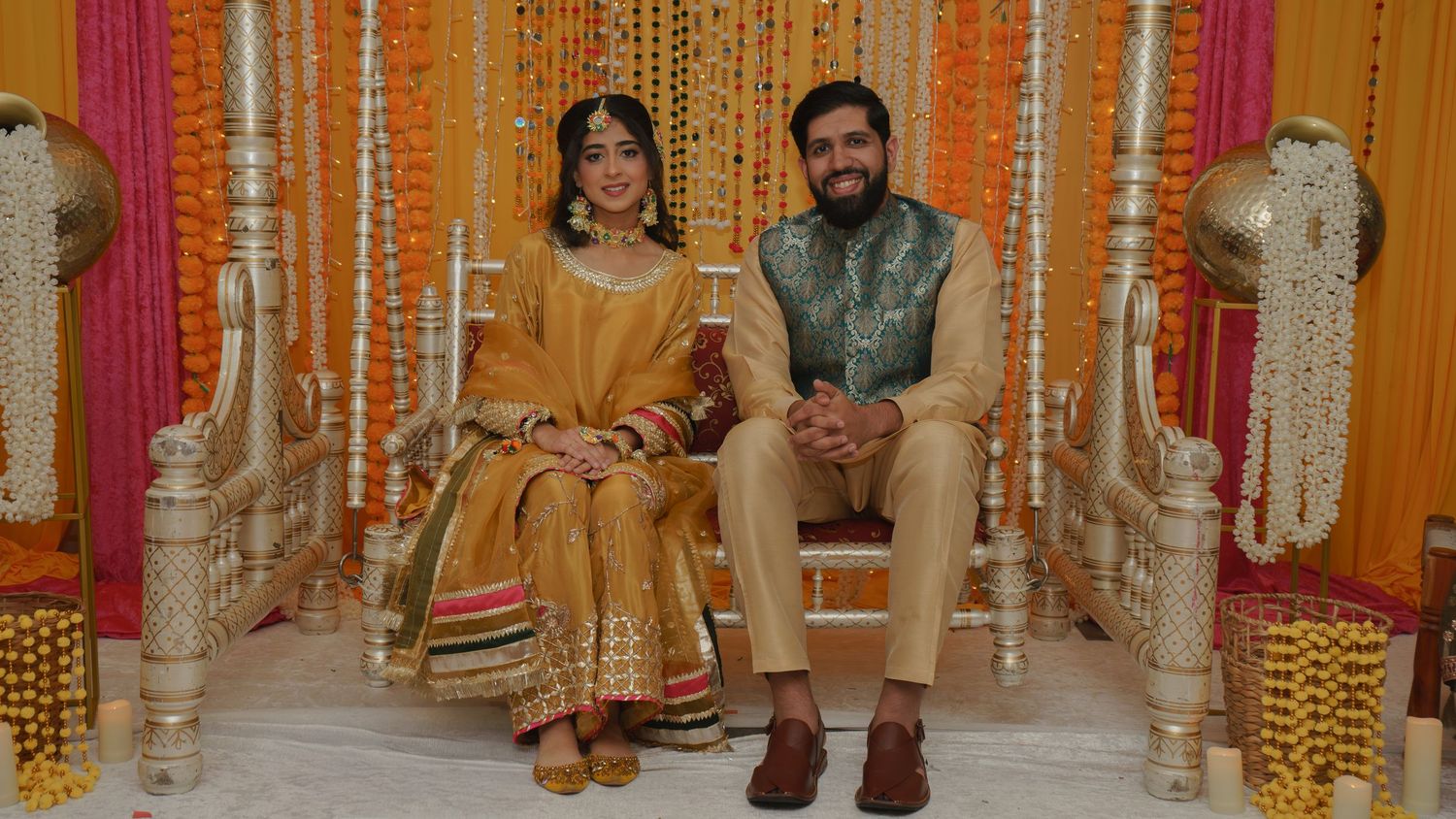 Couple in traditional South Asian wedding attire seated on ornate chairs with floral decorations.