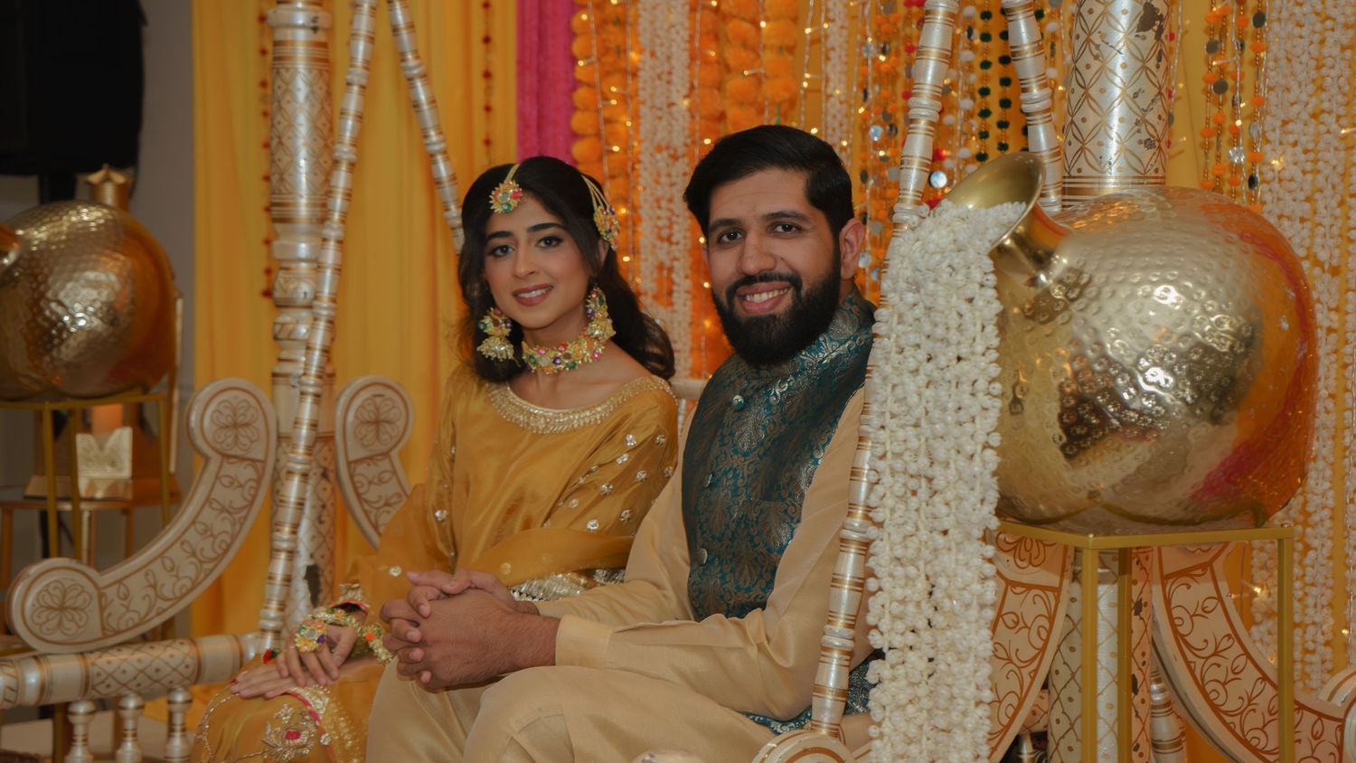 Couple in traditional South Asian wedding attire seated together, woman in gold saree, man in teal sherwani, floral decor.