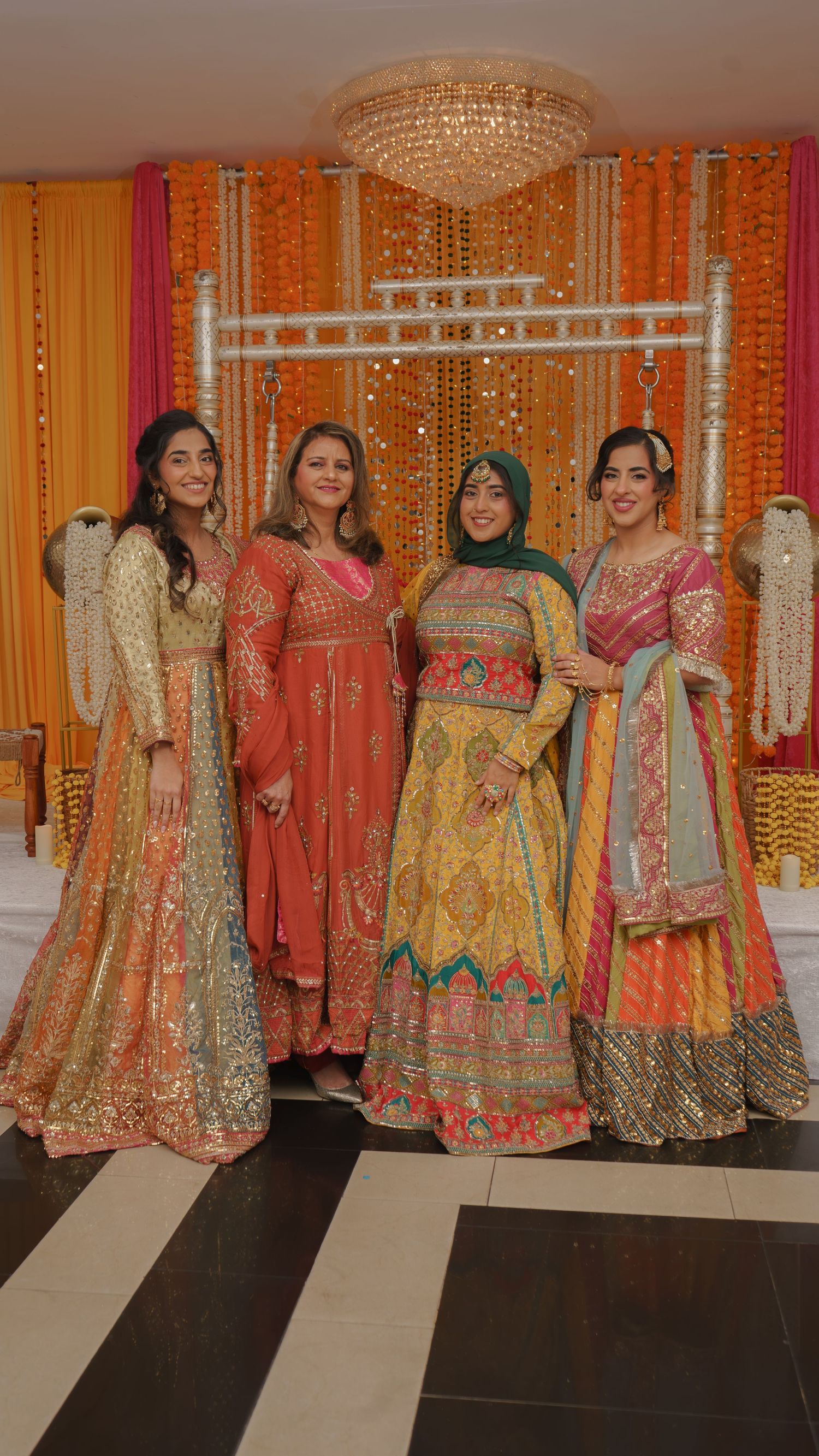 Four women in vibrant traditional South Asian attire pose together at a festive mehndi celebration with marigold decor.