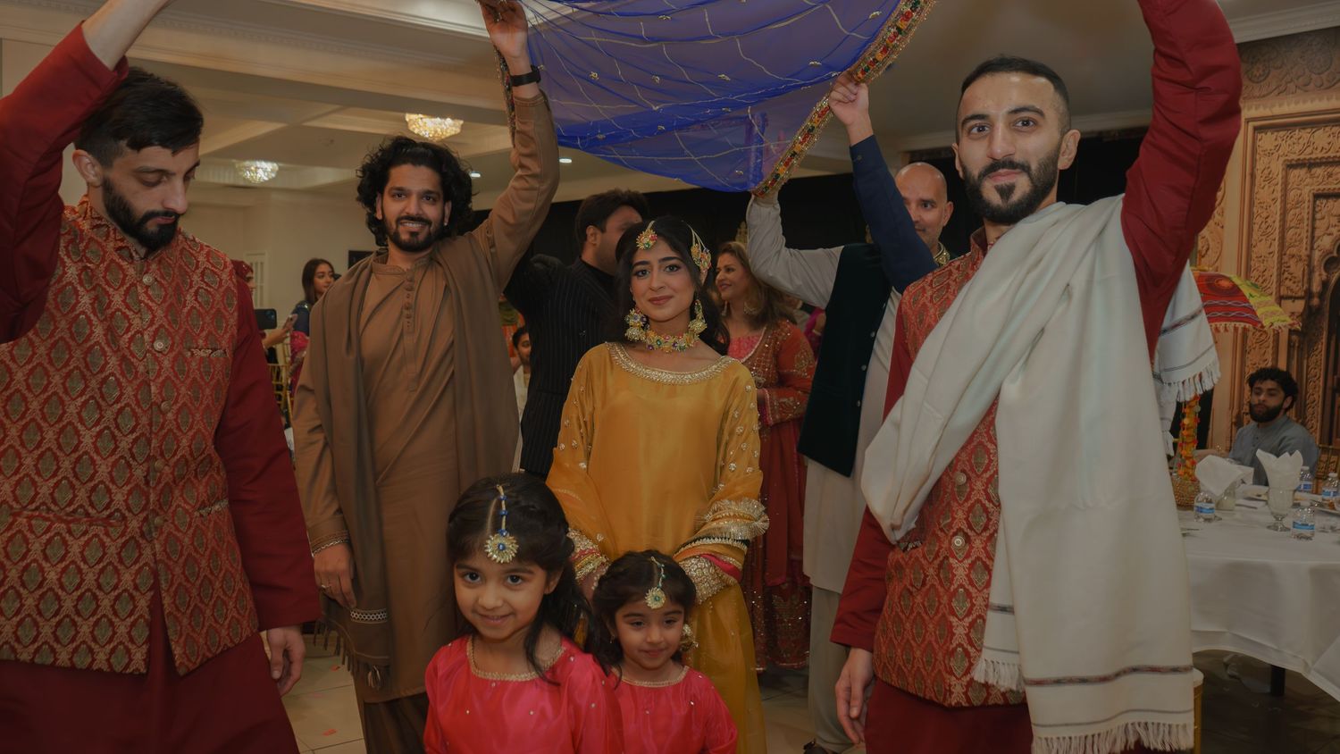 South Asian family in traditional attire celebrating at a festive gathering, holding a blue cloth overhead.