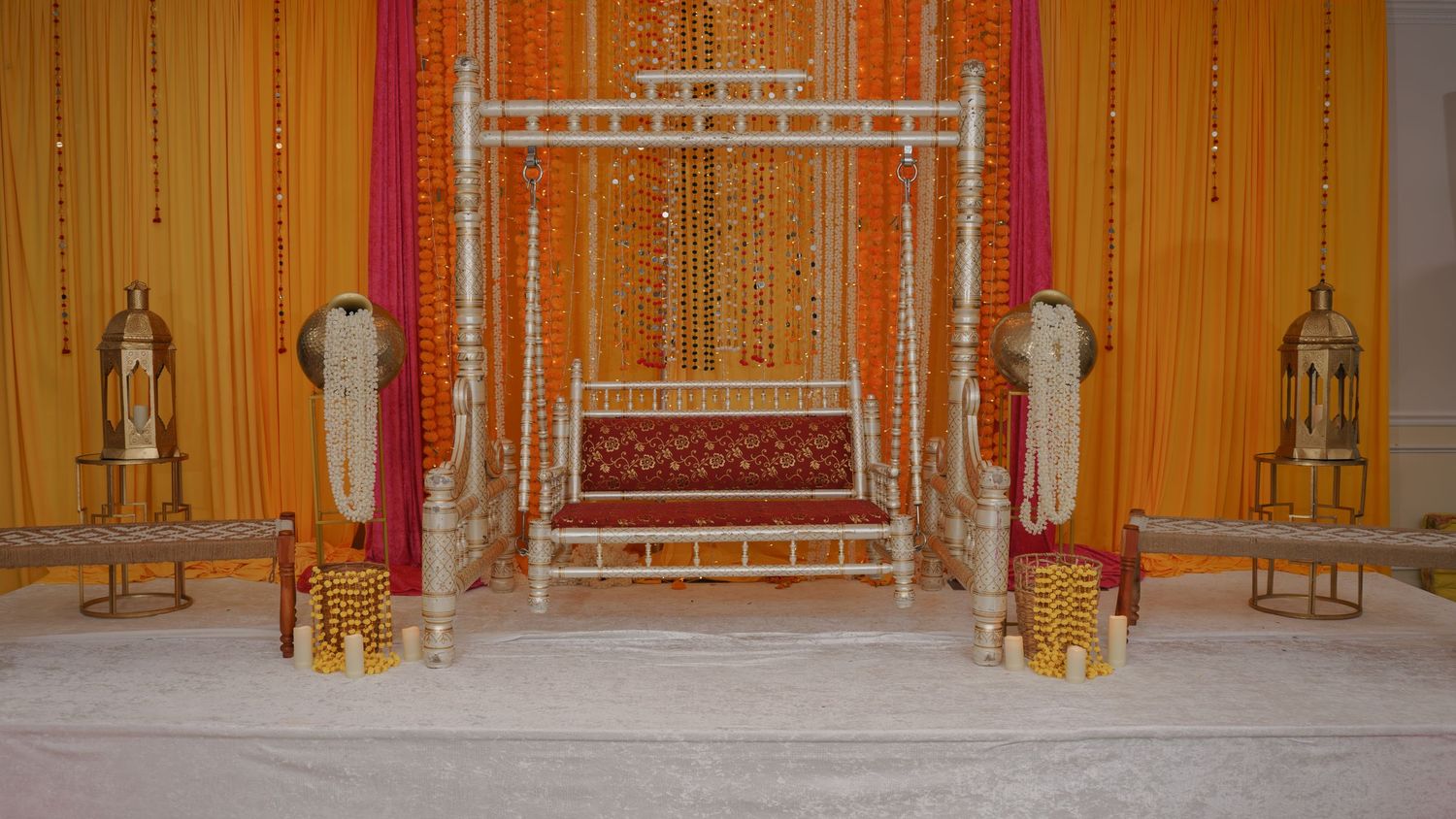 Elegant Indian wedding mandap with white floral arch, red velvet seating, orange drapes, and decorative lanterns.