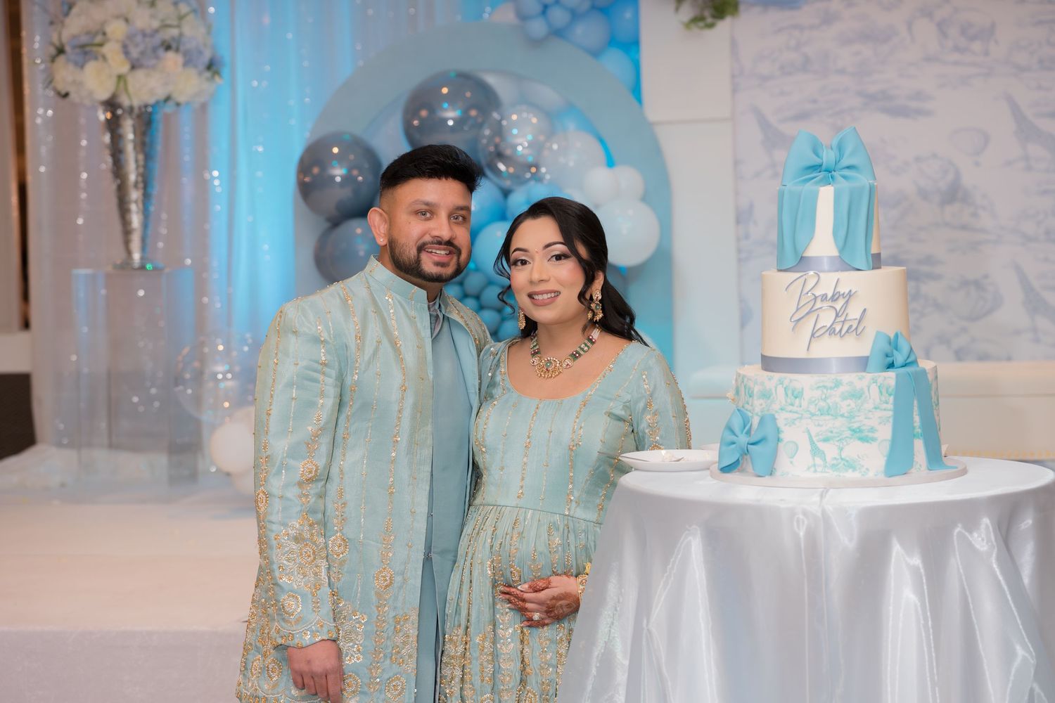 Couple in matching teal embroidered outfits at baby shower posing beside decorated gift-themed cake.