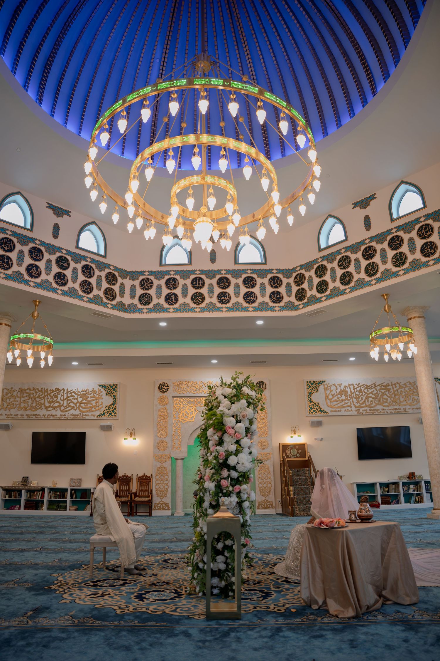 Elegant mosque interior with ornate chandelier, floral arch, and blue domed ceiling set for a wedding ceremony.