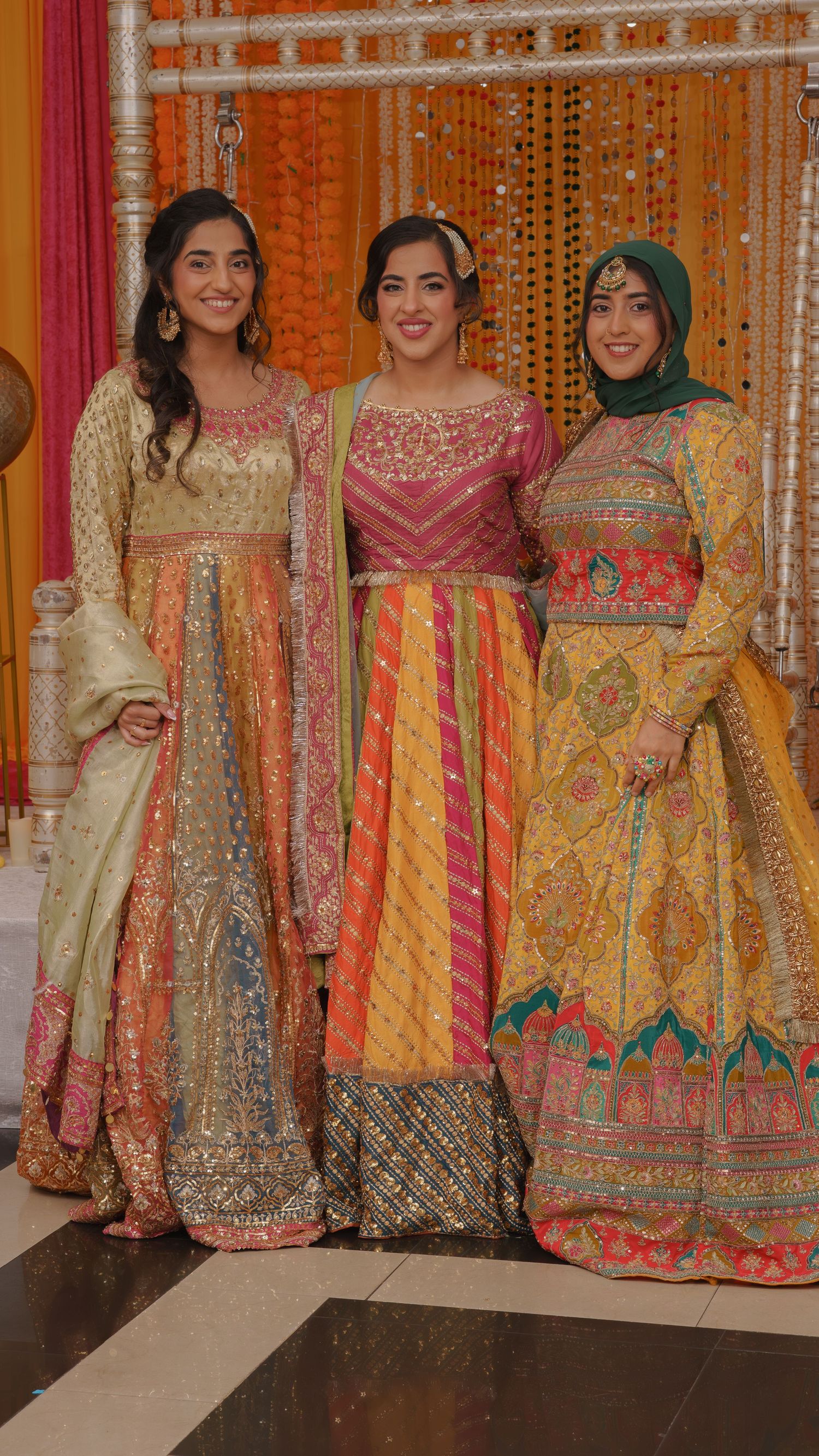 Three women in vibrant traditional South Asian attire smile together at a festive celebration with marigold decorations.