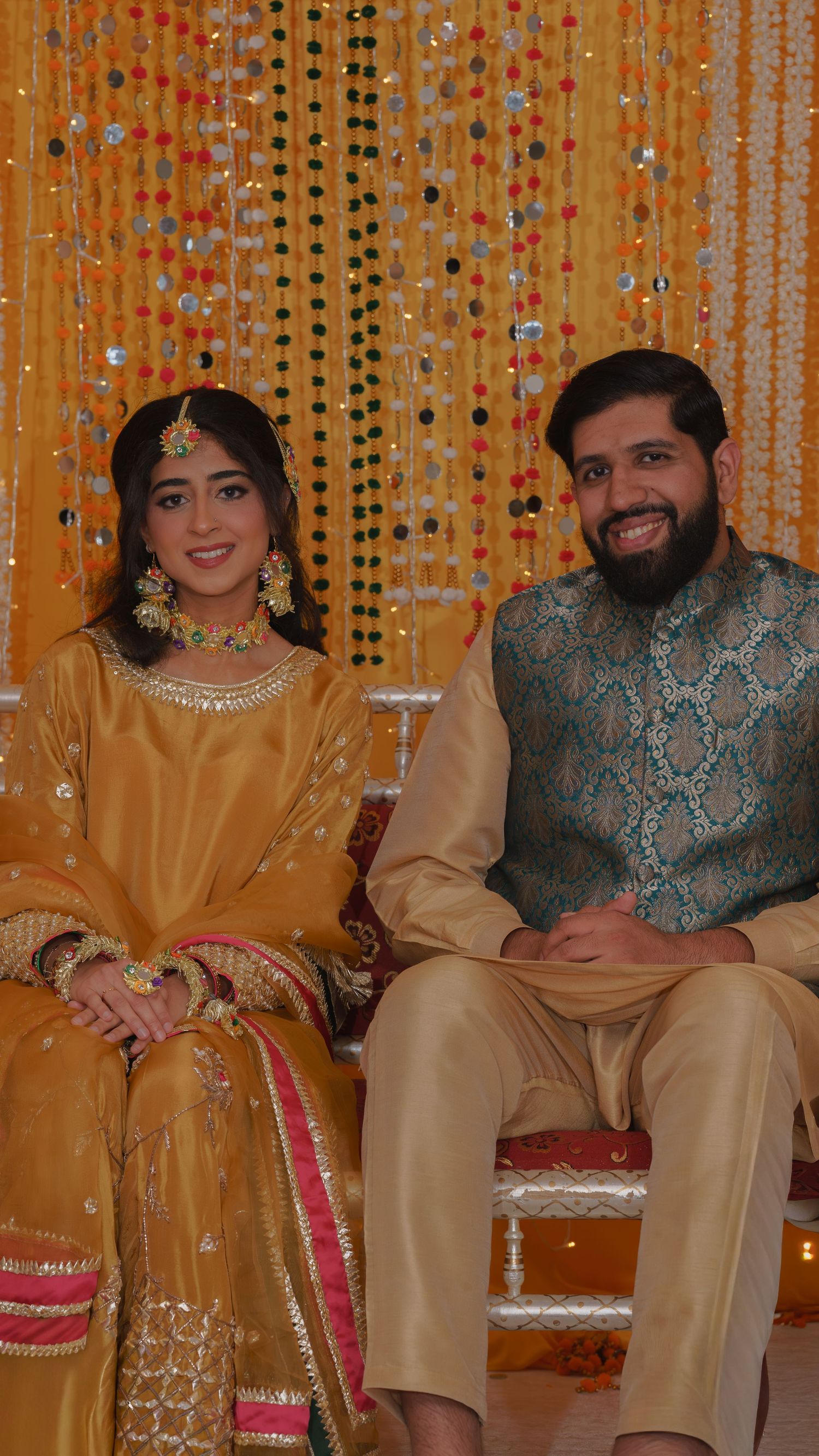 A couple in traditional South Asian attire sitting together at a festive celebration with colorful decorations.