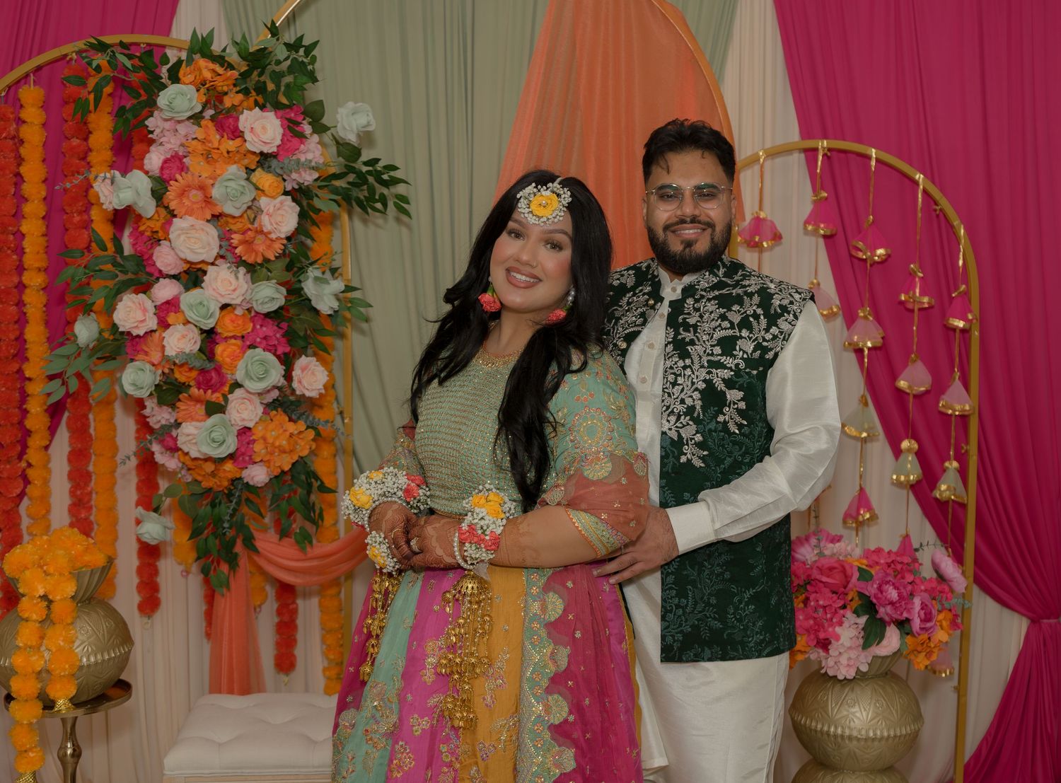 Couple in traditional South Asian attire posing at a colorful floral decorated Mehndi ceremony backdrop.