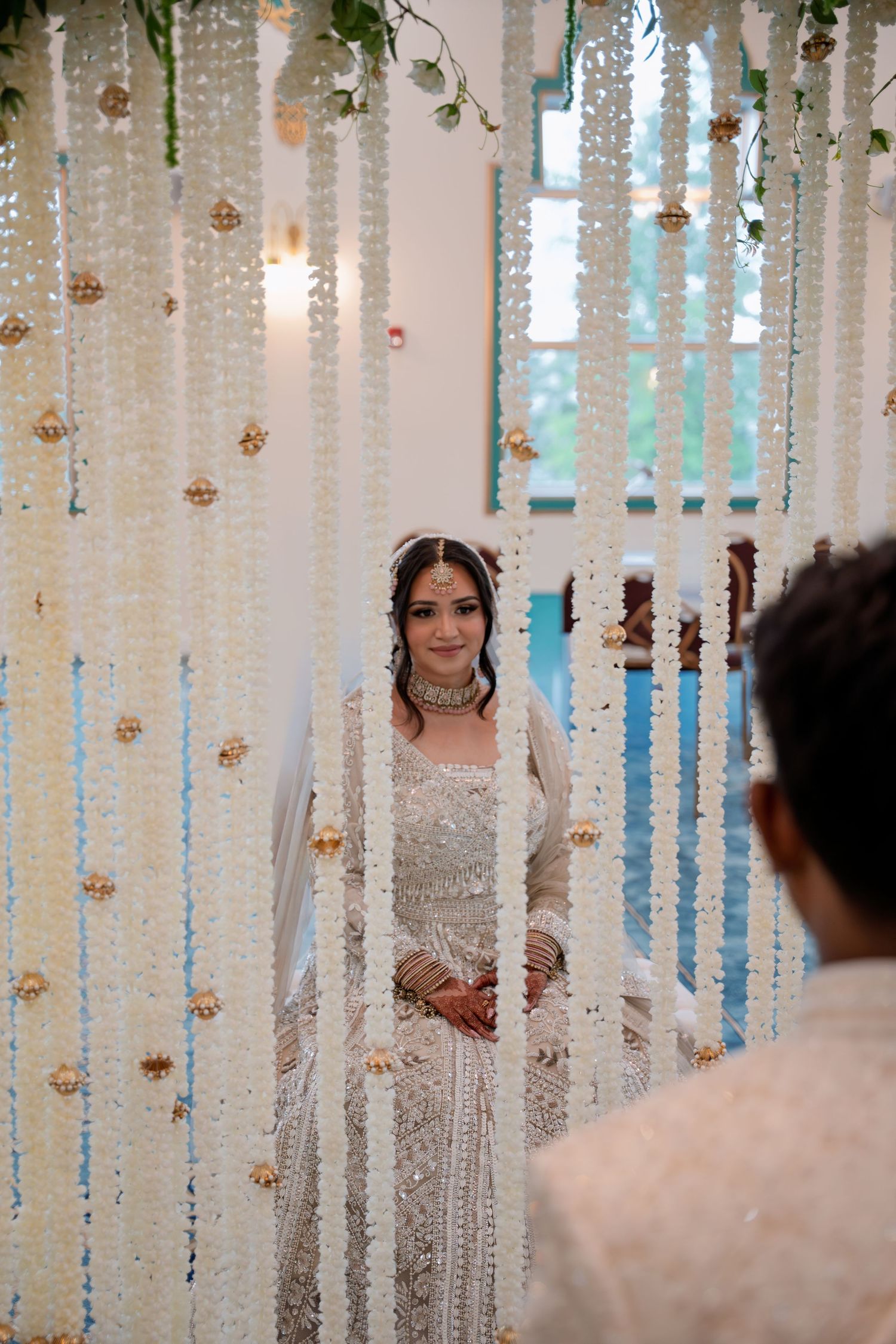 Bride in white embroidered lehenga stands behind floral curtain of white hanging garlands at Indian wedding ceremony.