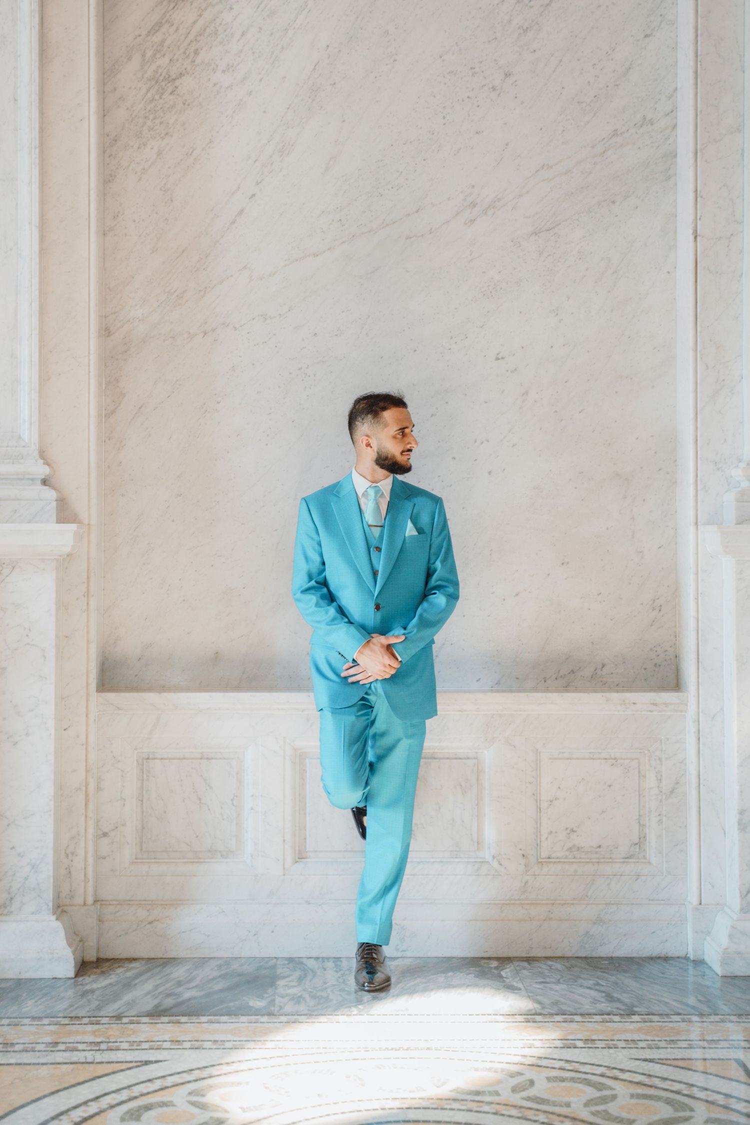 Confident man in turquoise suit leaning against marble wall in grand interior.