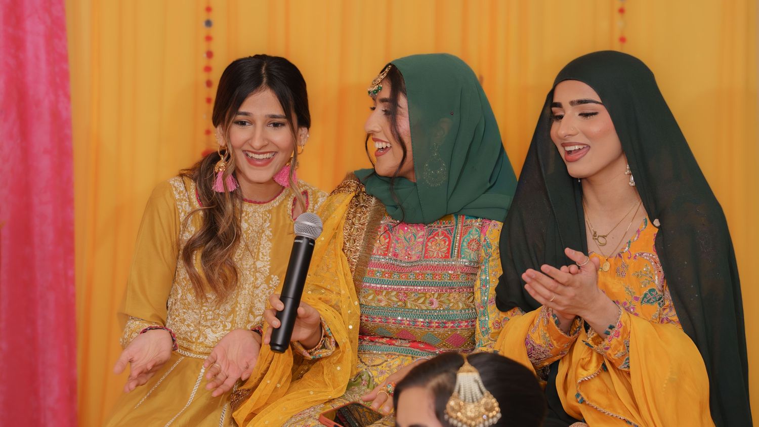 Three women in colorful traditional attire smile and laugh together at a festive cultural celebration.