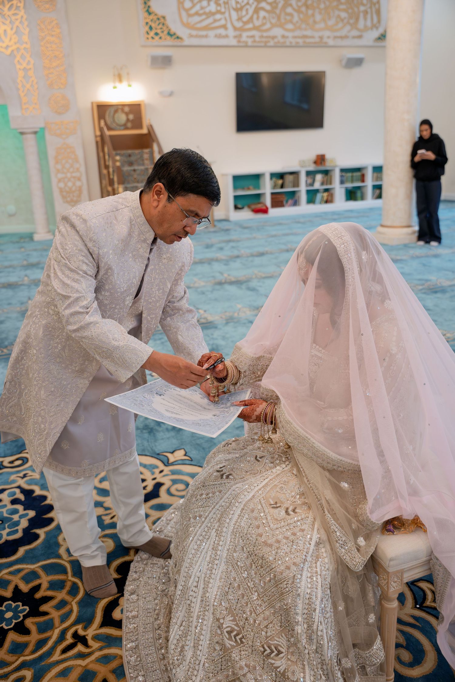 Groom in white sherwani signs nikah marriage contract with veiled bride in ornate mosque ceremony.