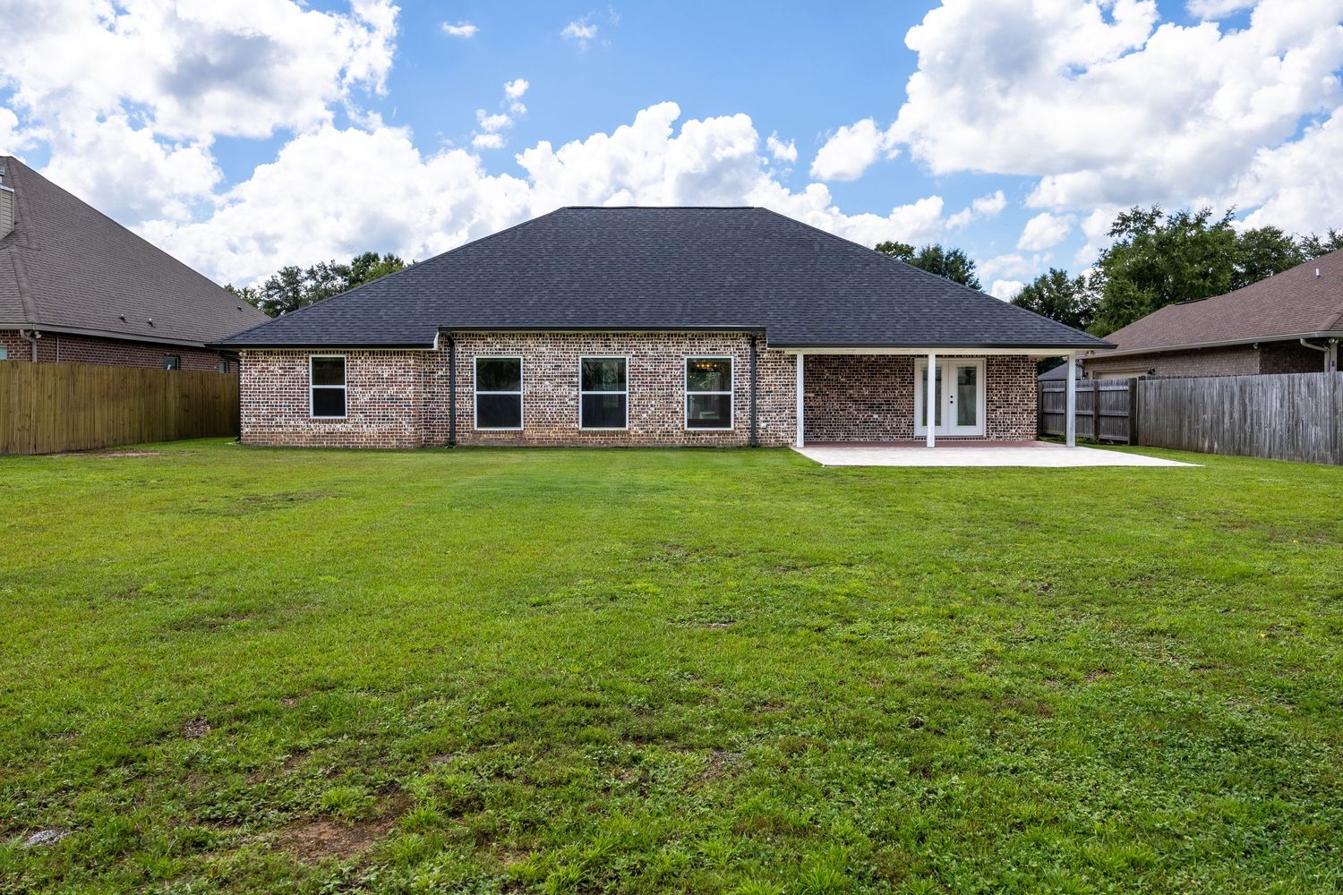 Brick ranch-style home with dark roof, large green backyard, patio, and wood privacy fence on a sunny day.