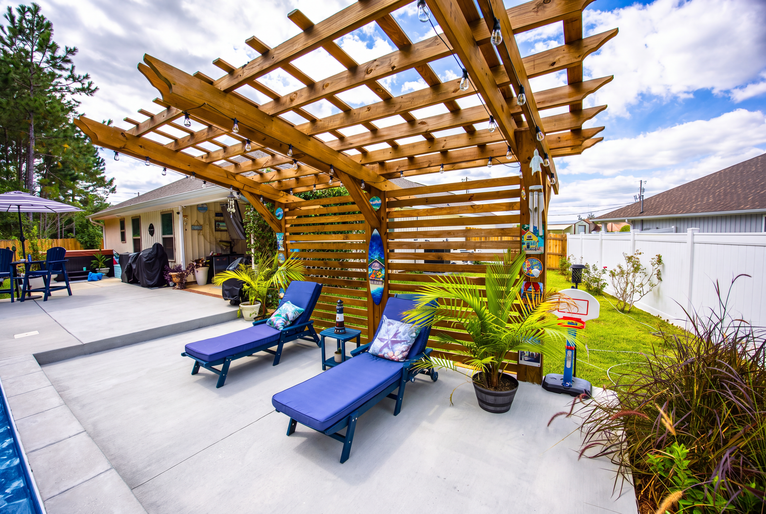 Outdoor patio with wooden pergola, blue lounge chairs, and pool area in a sunny backyard setting.