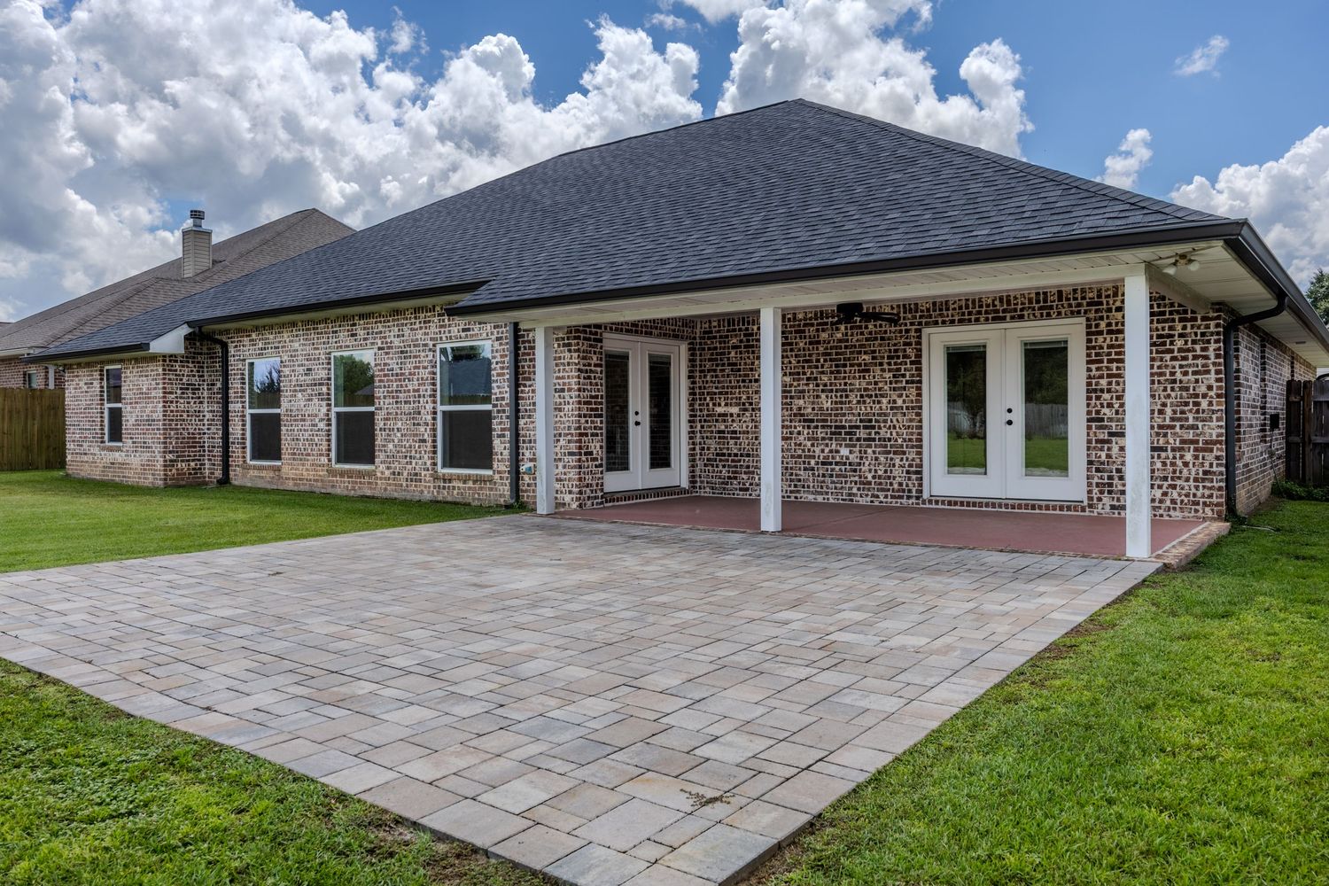 Brick home exterior with covered patio, French doors, and decorative stone pavers surrounded by lush green lawn.