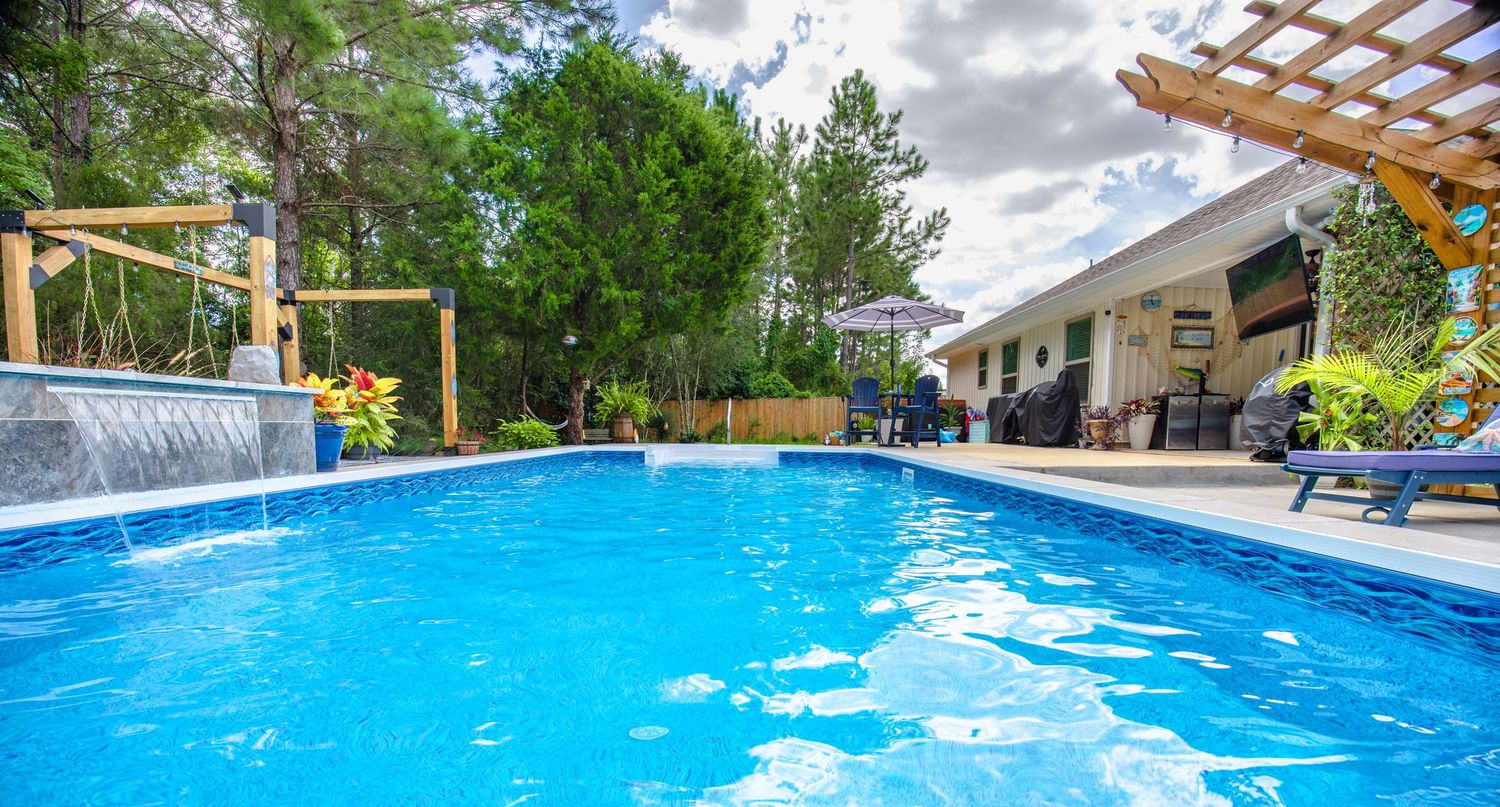 Sparkling blue swimming pool with waterfall feature, pergola, and lush trees in a beautiful backyard oasis.