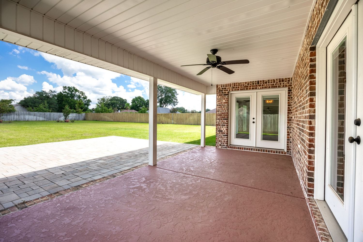 Covered brick patio with ceiling fan and French doors leading to a spacious green backyard on a sunny day.