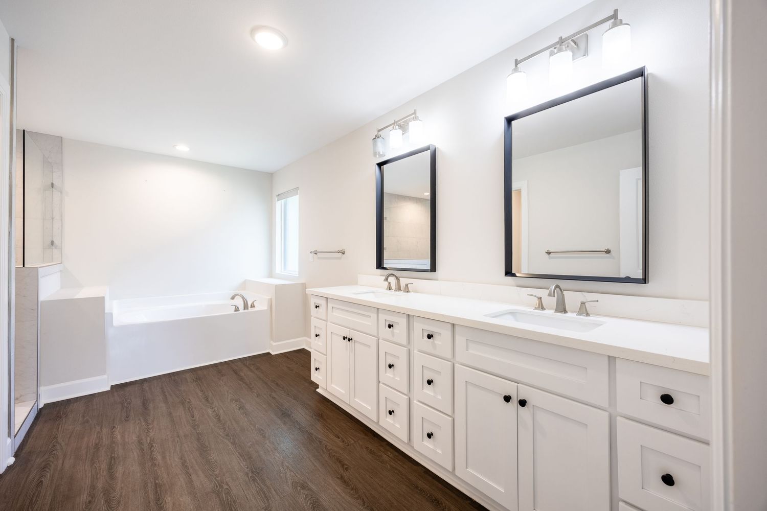 Bright white bathroom with double vanity, dark wood floors, dual mirrors, and a soaking tub near a window.