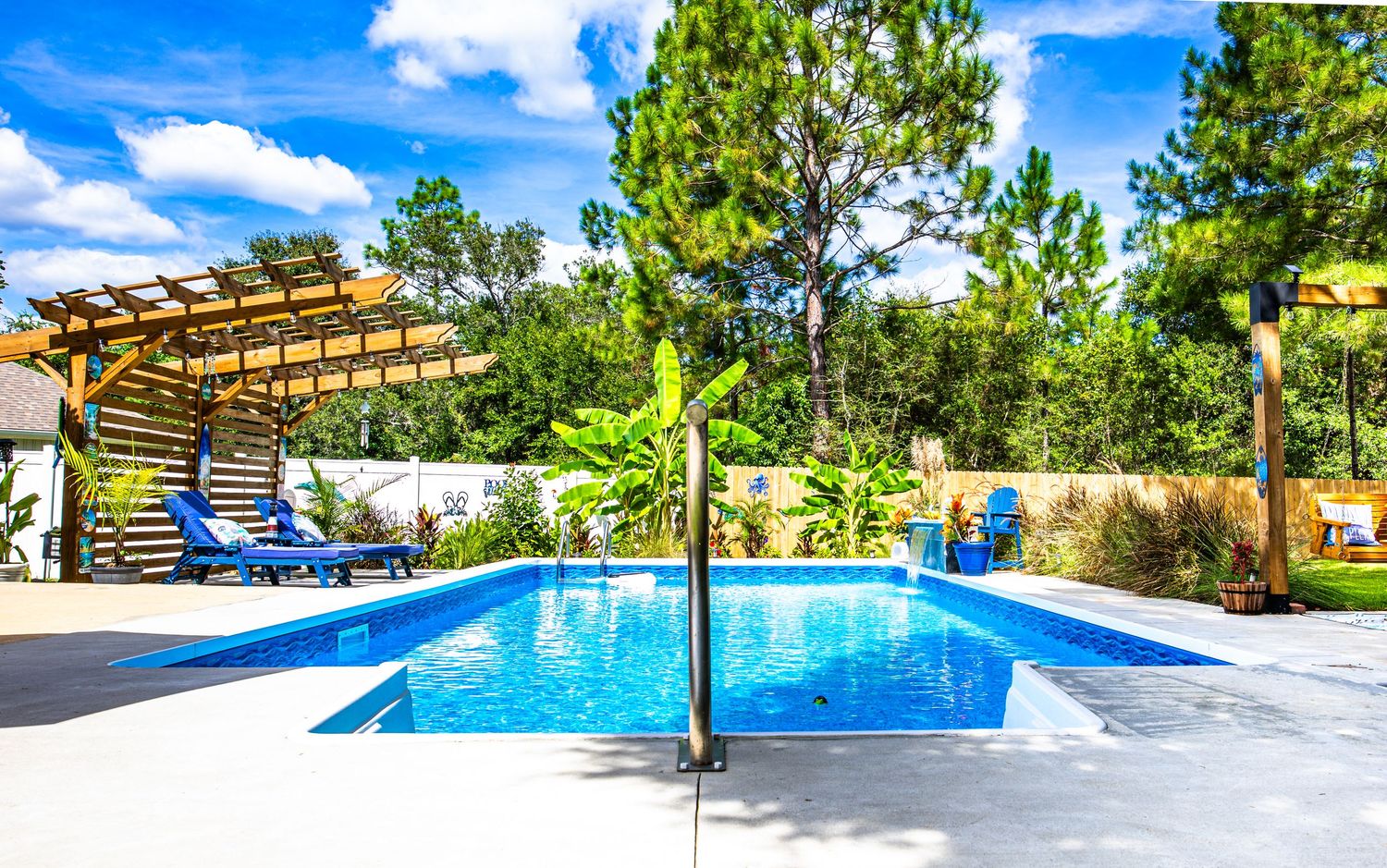 Outdoor swimming pool with blue lounge chairs, wooden pergola, and tropical landscaping under a bright blue sky.