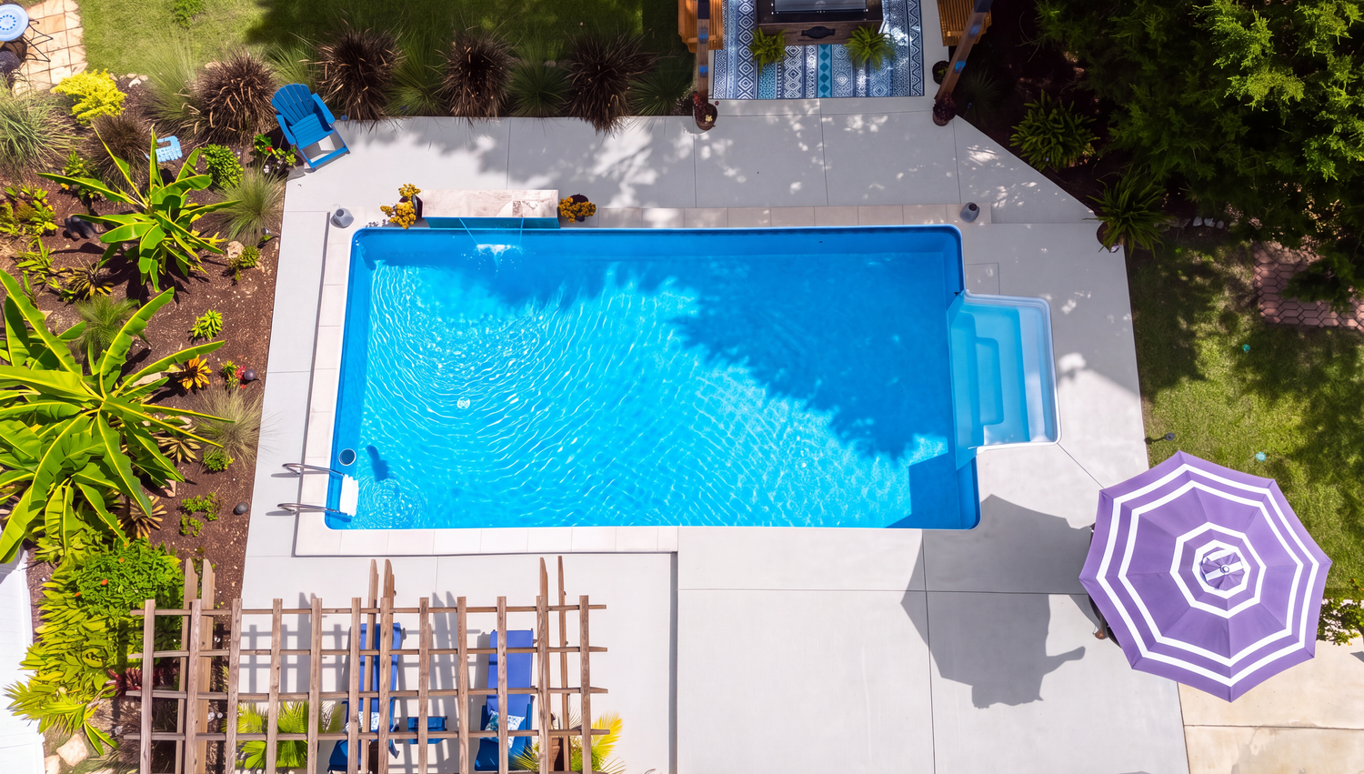 Aerial view of a rectangular blue swimming pool surrounded by a white concrete deck, lush greenery, and a purple striped umbrella.