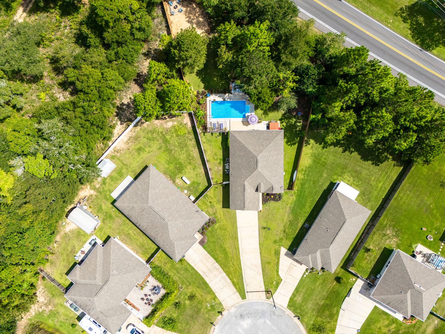 Aerial drone view of suburban homes with lush green trees, manicured lawns, and a backyard swimming pool near a road.