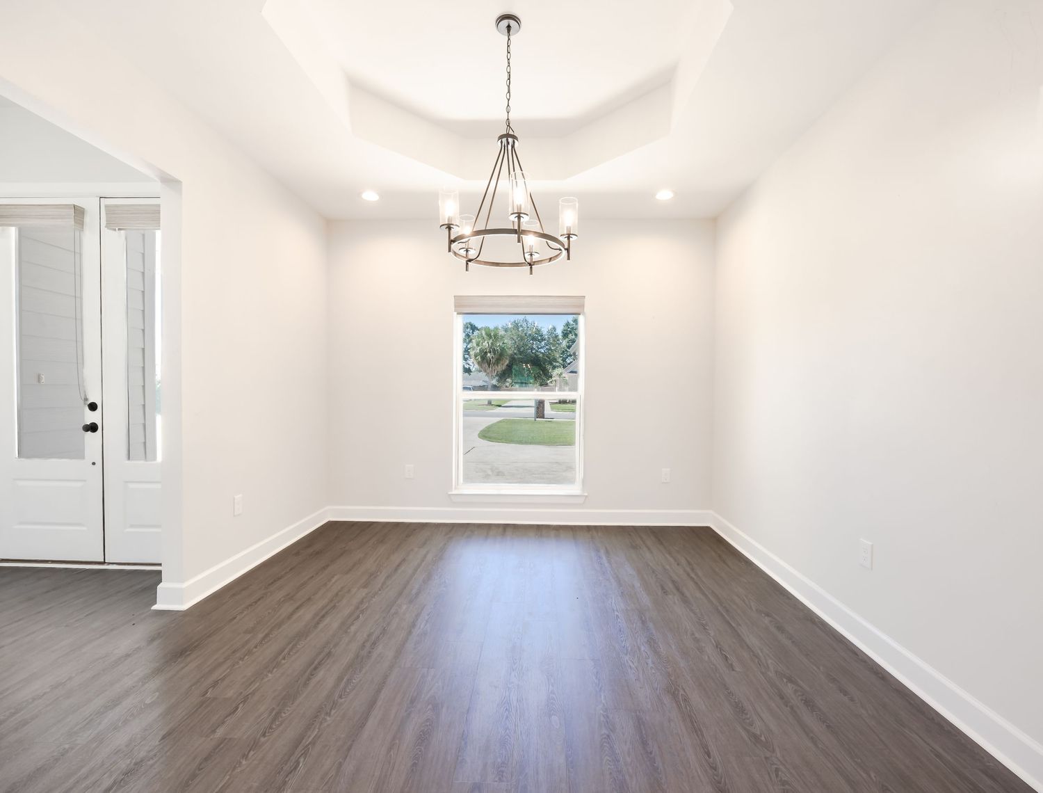 Bright empty dining room with tray ceiling, chandelier, dark hardwood floors, and large window.