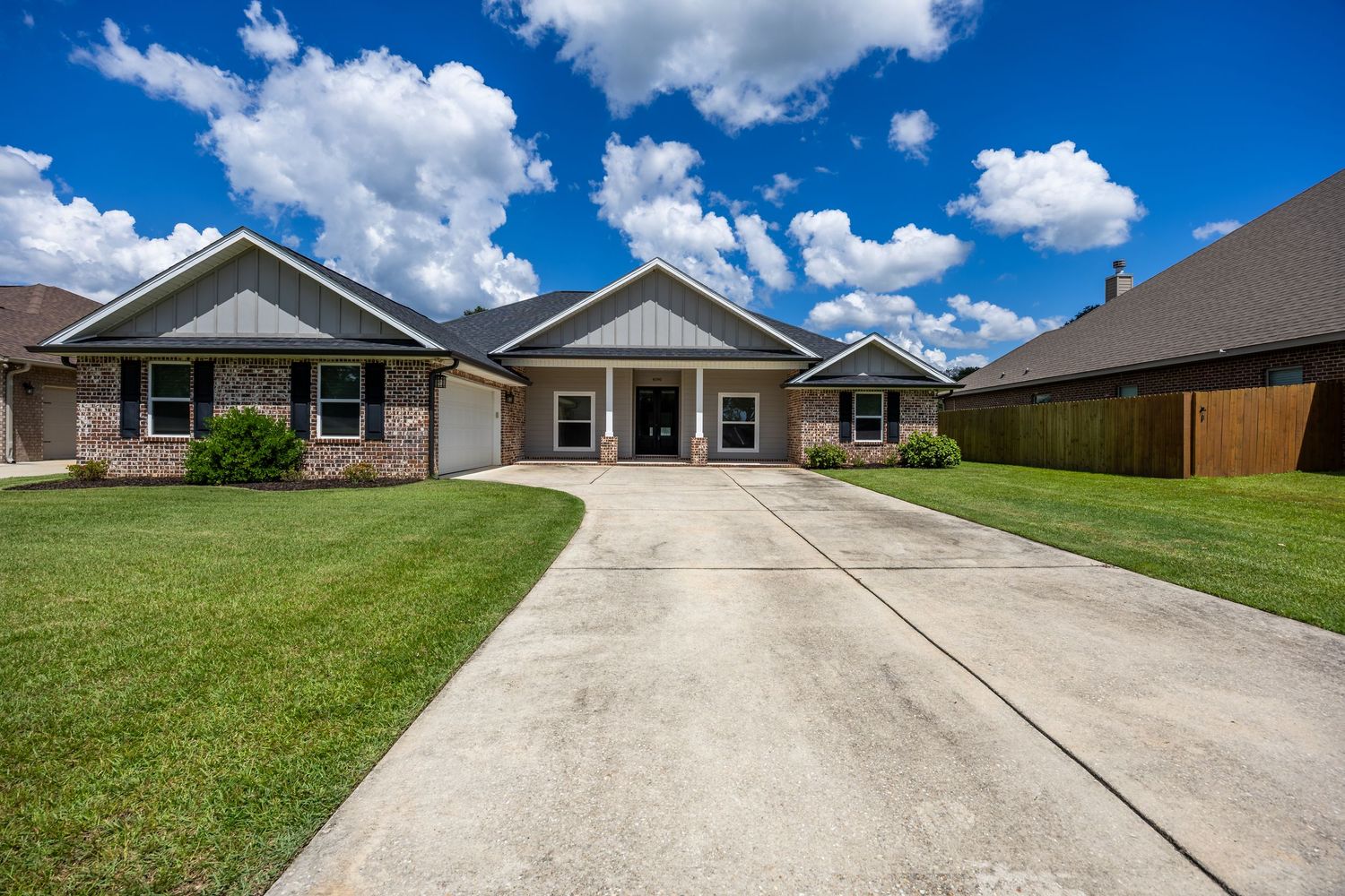 Single-story brick and board-and-batten home with wide driveway, attached garage, and lush green lawn under blue skies.