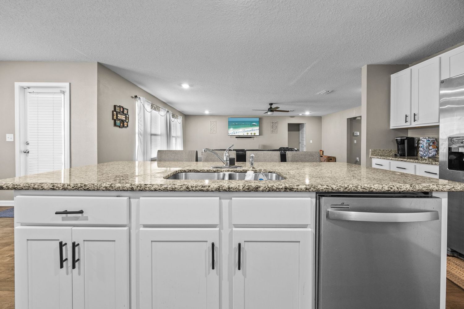 Modern kitchen with white cabinets, granite countertops, stainless steel dishwasher, and open-concept living area in background.