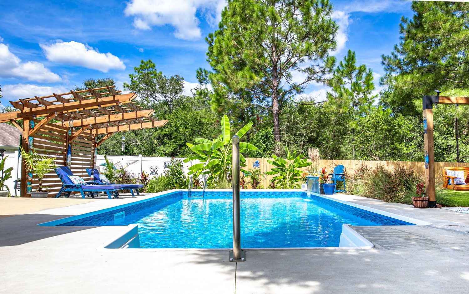 Luxury outdoor pool with pergola, lounge chairs, and lush green trees under a blue sky.