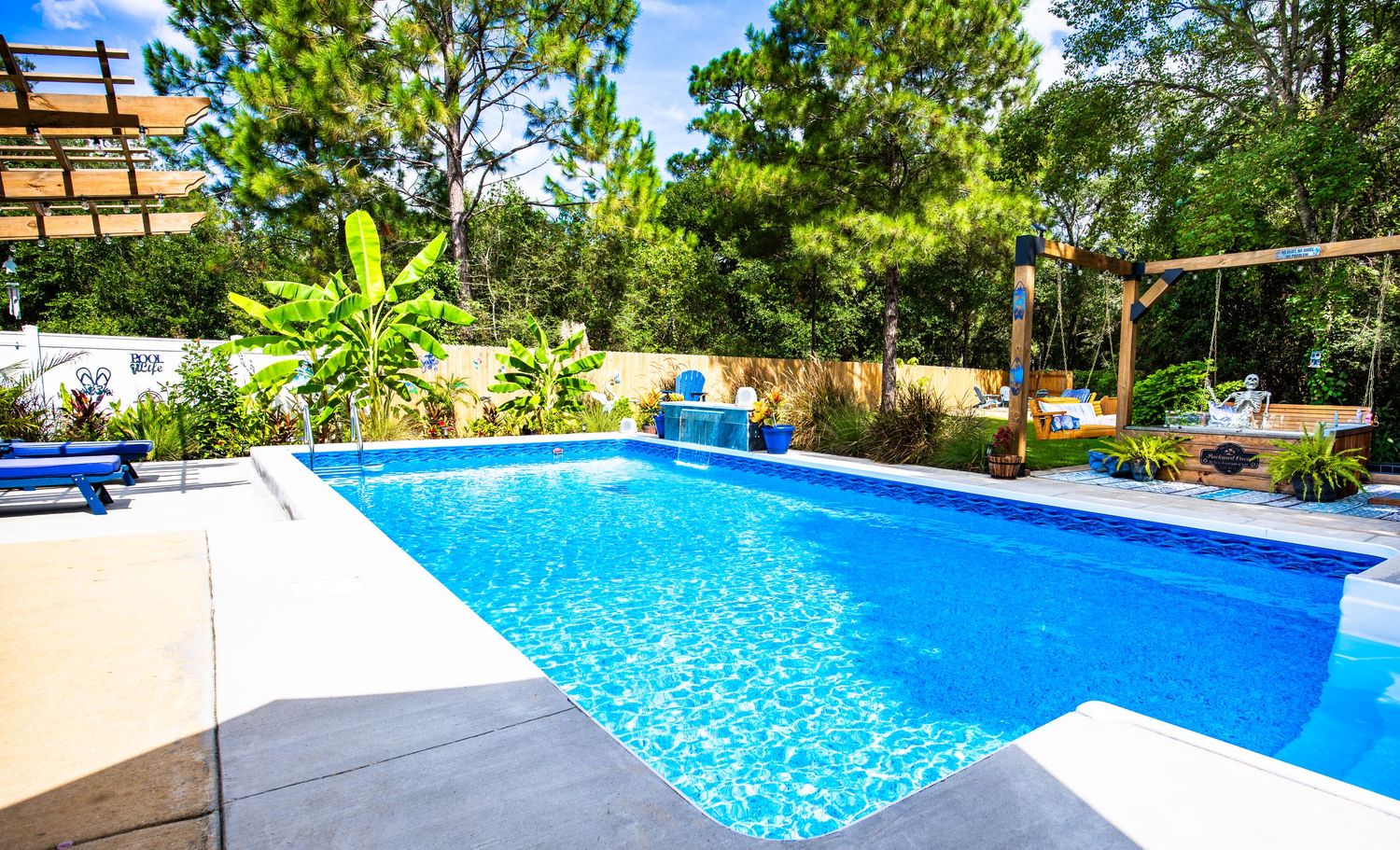 Luxury outdoor swimming pool with crystal blue water surrounded by lush tropical landscaping and wooden pergola on sunny day.