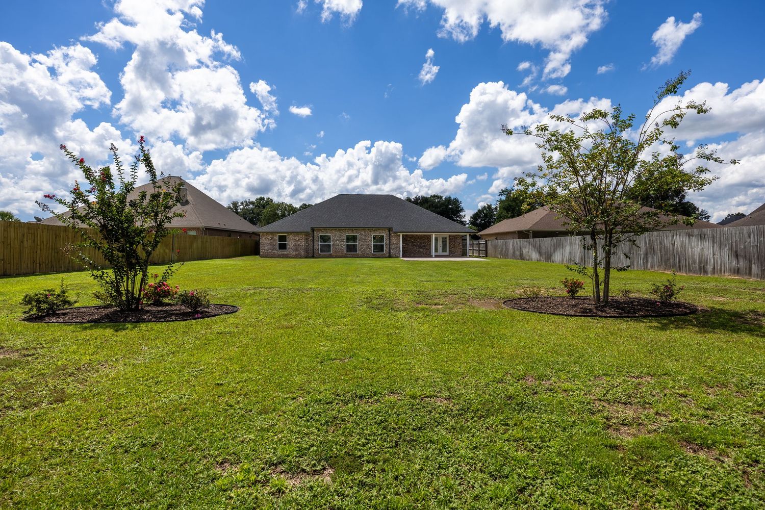 Spacious backyard with lush green lawn, mature trees, wood privacy fence, and single-story home under blue sky.