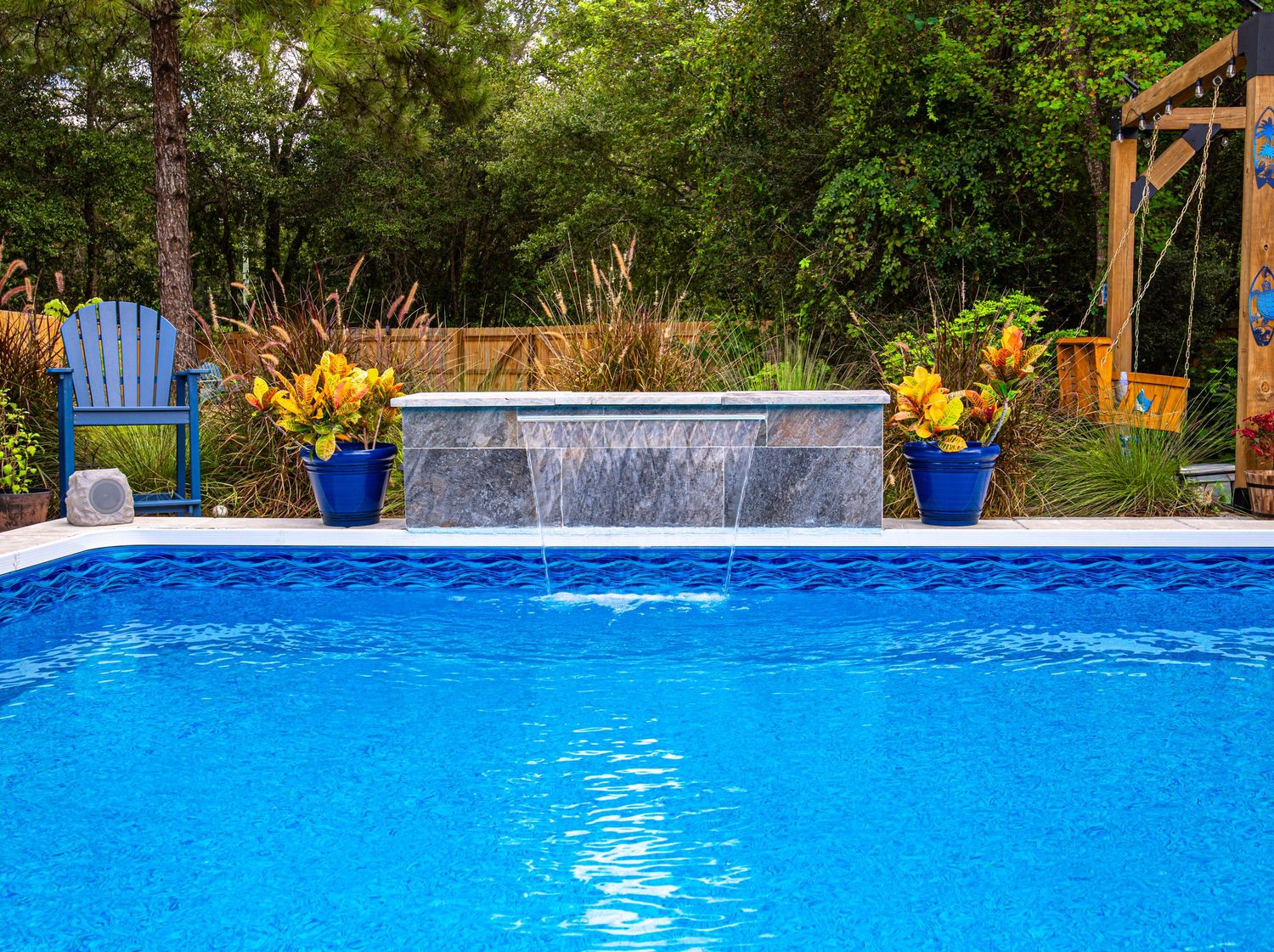 Blue backyard pool with waterfall feature, potted yellow flowers, and lush green trees in background.