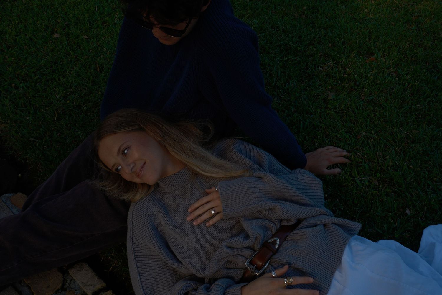 A woman with long blonde hair rests her head on a man's lap on a dark grassy lawn at dusk, smiling softly.