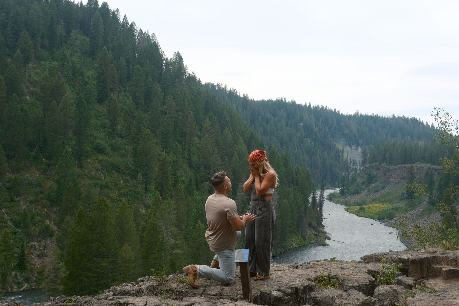 A man proposes on one knee to a woman in a red beanie at a cliffside viewpoint above a river and pine-covered hills.