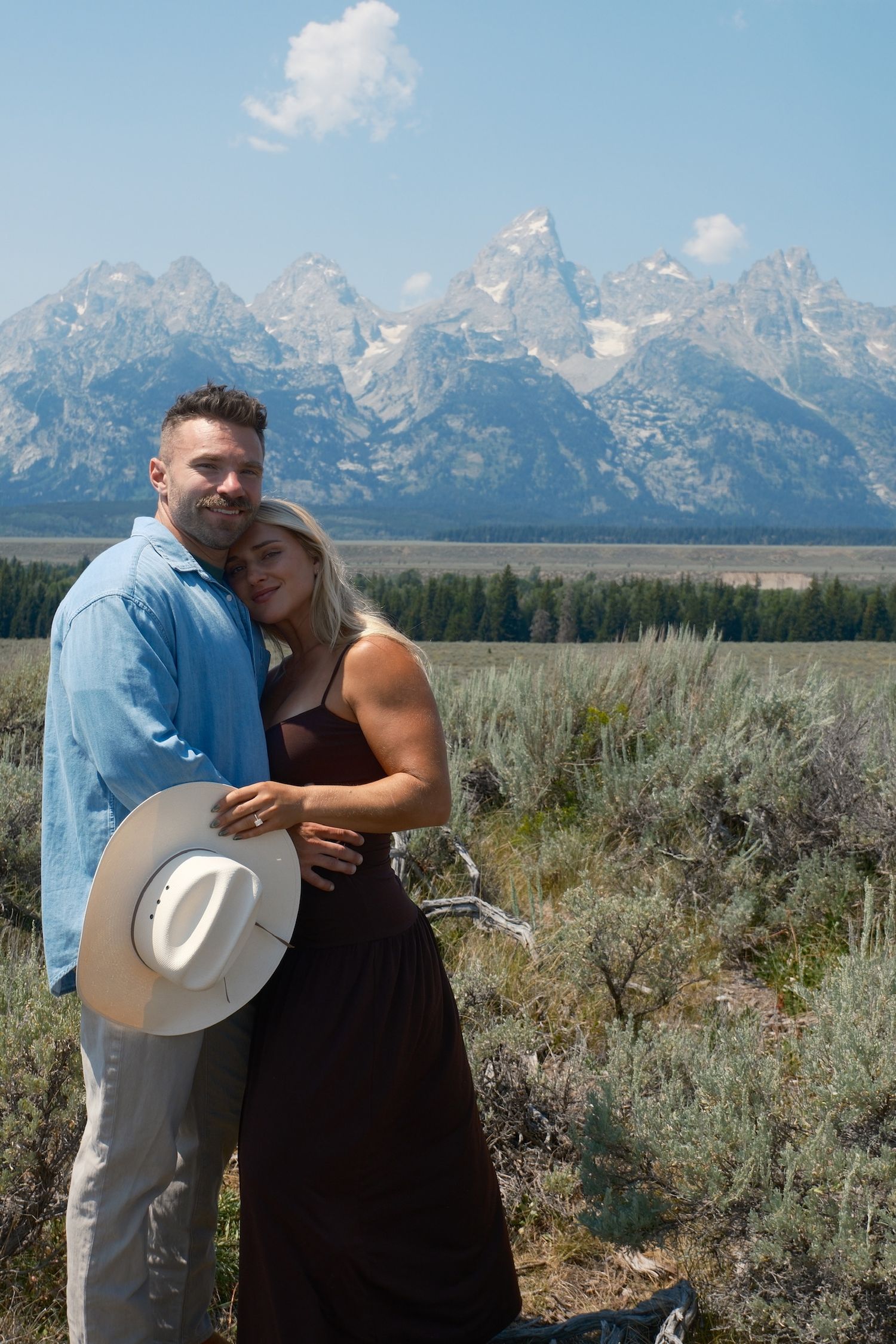 Smiling couple hugging in a sagebrush meadow, the man holding a white cowboy hat with mountains in the background.