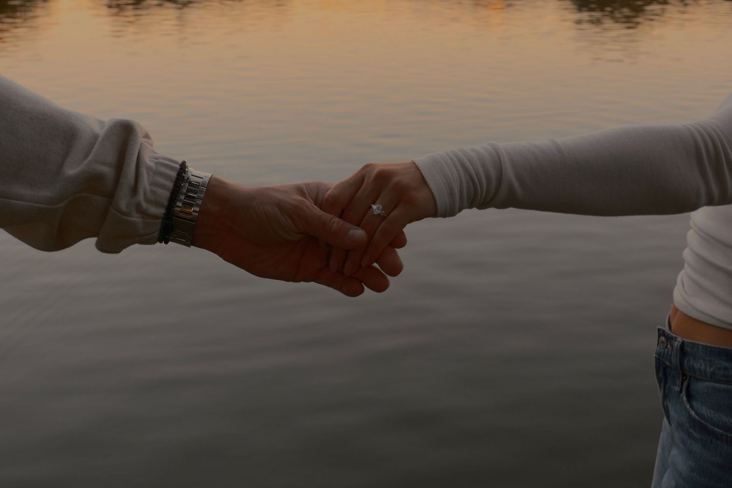 Two people hold hands over calm water at sunset, one wearing a watch and the other a ring.